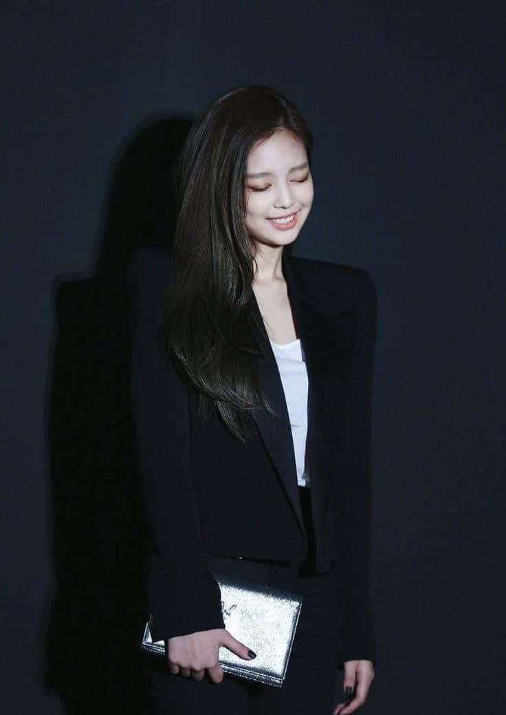 Jennie Kim 桌布 壁紙 — 一位身穿黑色西裝外套和白色上衣的女性，在深色背景下輕輕微笑，手持銀色手拿包。