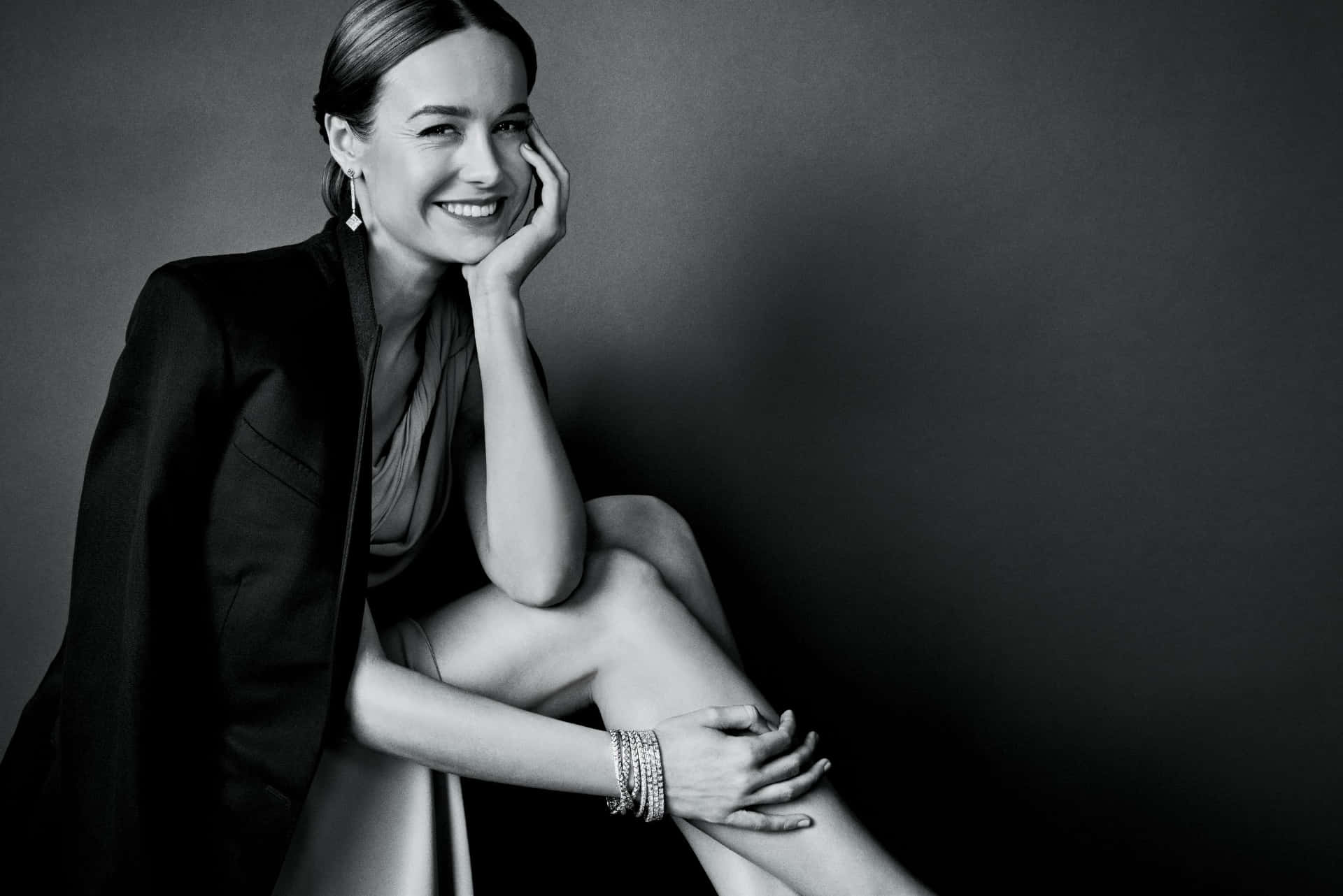 brie larson 桌布 壁紙 — 一位身穿黑色西裝外套的女性，在深灰色工作室背景前，將下巴擱在手上，露出溫暖的笑容。