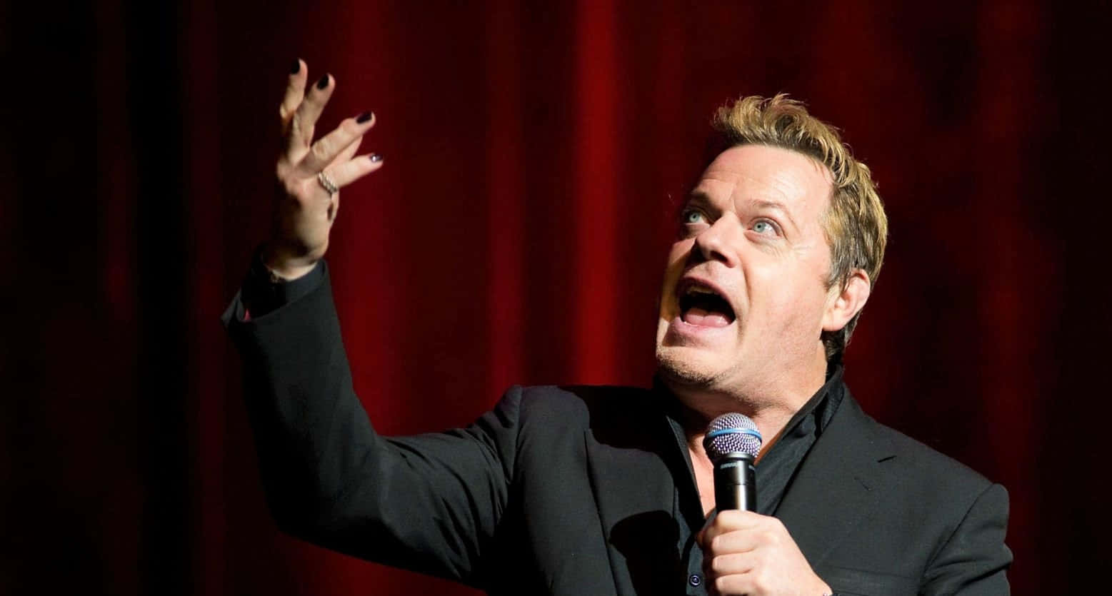 Eddie Izzard 桌布 壁紙 — 一位身穿黑色西裝的喜劇演員在舞台上表演，手持麥克風，舉起手，背景是紅色帷幕。