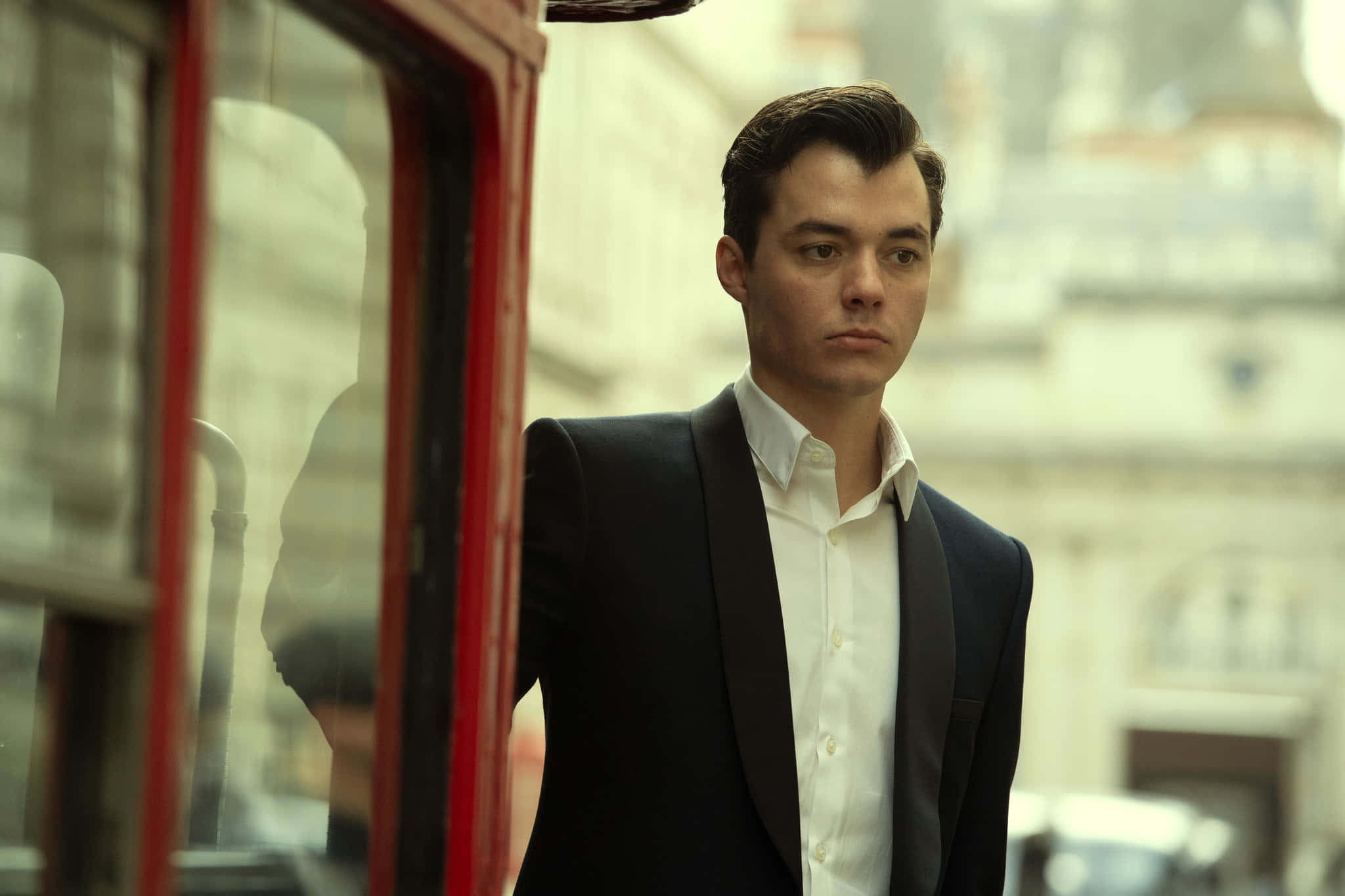 Alfred Pennyworth 桌布 壁紙 — 一位身穿黑色西裝的年輕男子站在城市街道上的紅色電話亭旁，背景是模糊的歷史建築。