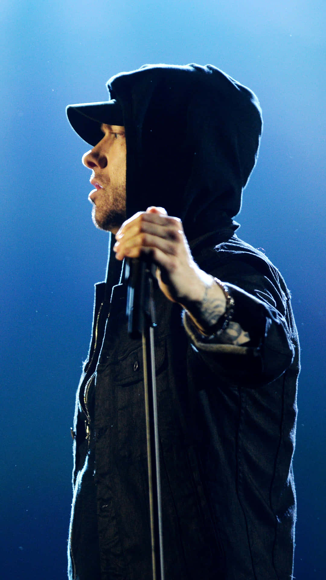 Eminem 桌布 壁紙 — 一位身穿黑色連帽衫和帽子的男性表演者在強烈的藍色舞台燈光下拿著麥克風。