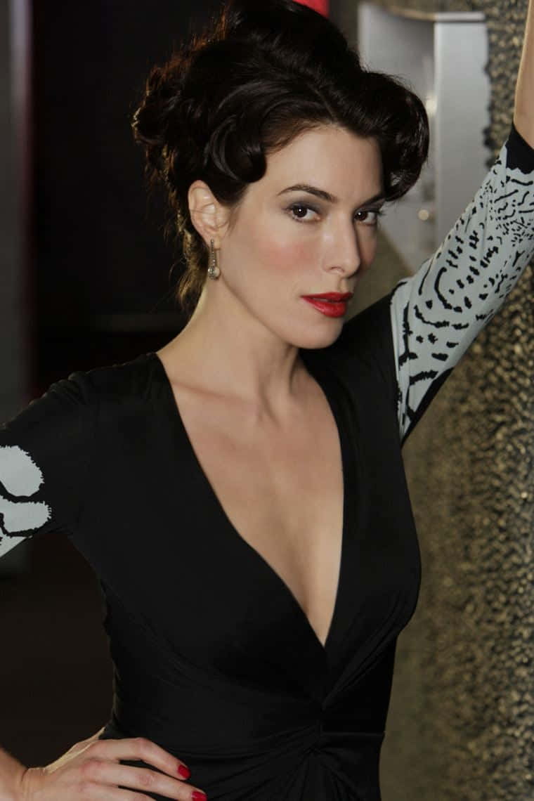 Jaime Murray 桌布 壁紙 — 一位身穿黑色連衣裙、袖子上有圖案的優雅女性，自信地靠在有紋理的牆上擺姿勢。