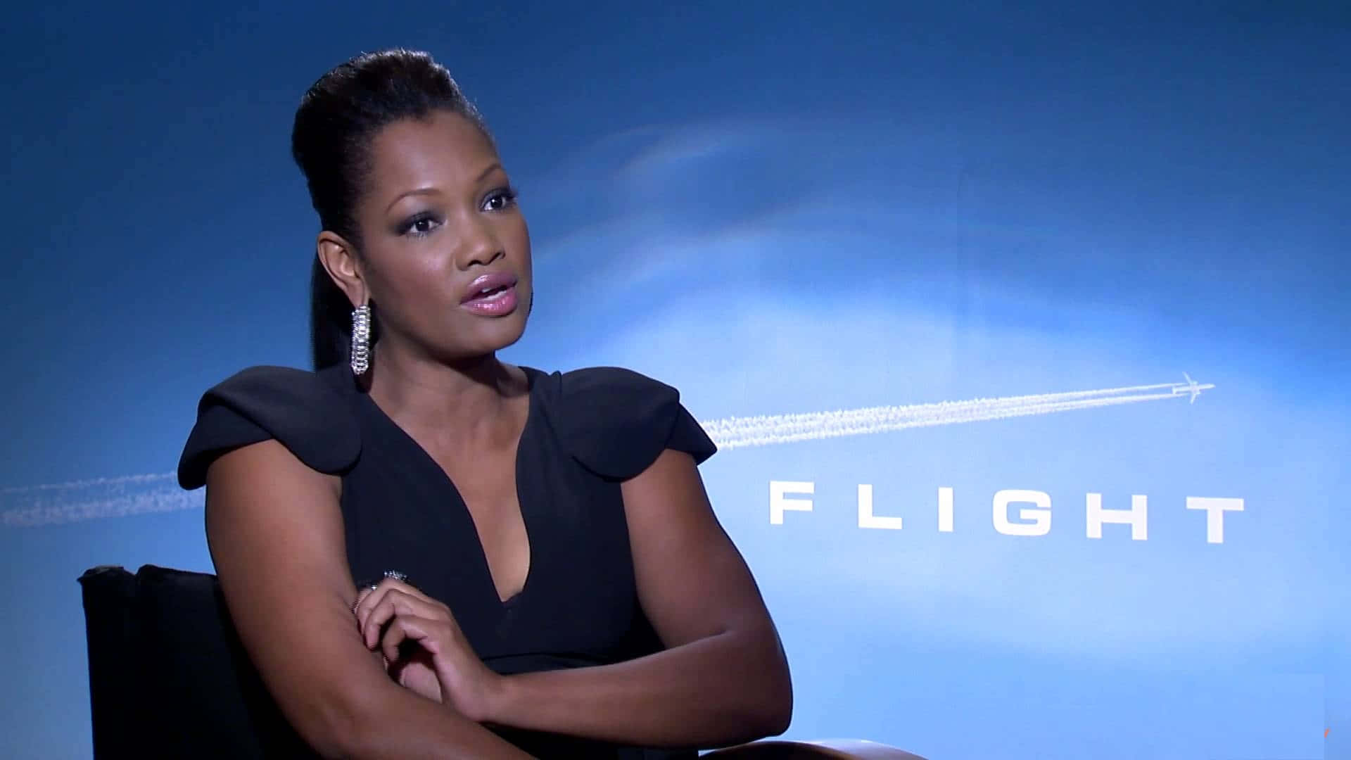 Garcelle Beauvais 桌布 壁紙 — 一位身穿黑色連衣裙的優雅女士坐在藍色背景前，背景上有「FLIGHT」字樣和飛機尾跡。