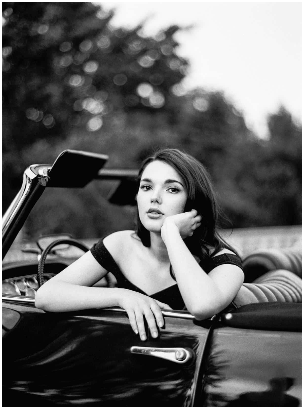 Black And White Vintage Car 桌布 壁紙 — 一位身穿黑色連衣裙的女性優雅地靠在一輛復古敞篷車的車門上，在柔和模糊的樹木背景下若有所思地凝視。