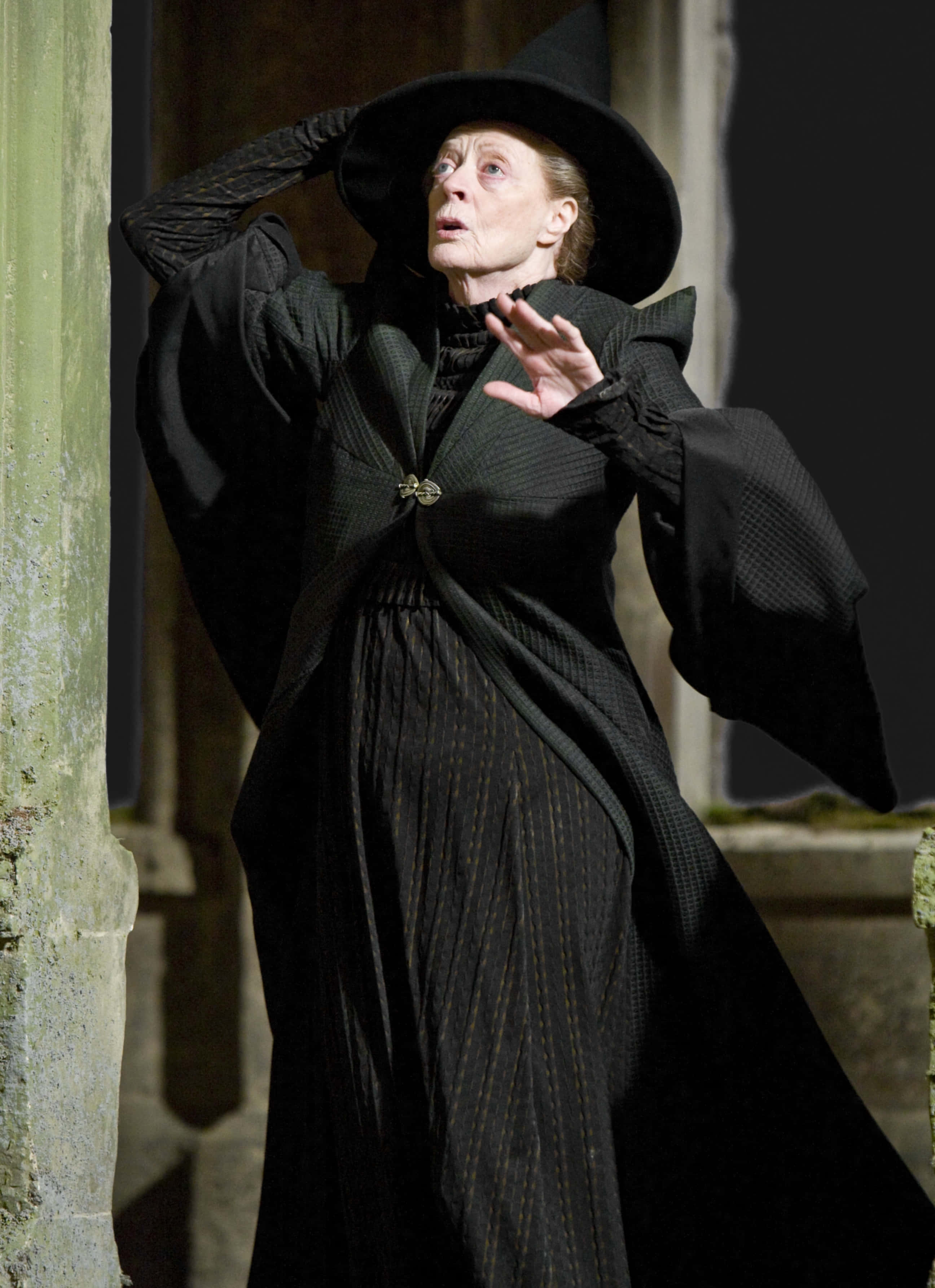 The Hogwarts Professors 桌布 壁紙 — 一位身穿黑色長袍和帽子的老女巫在石頭走廊中戲劇性地做手勢。