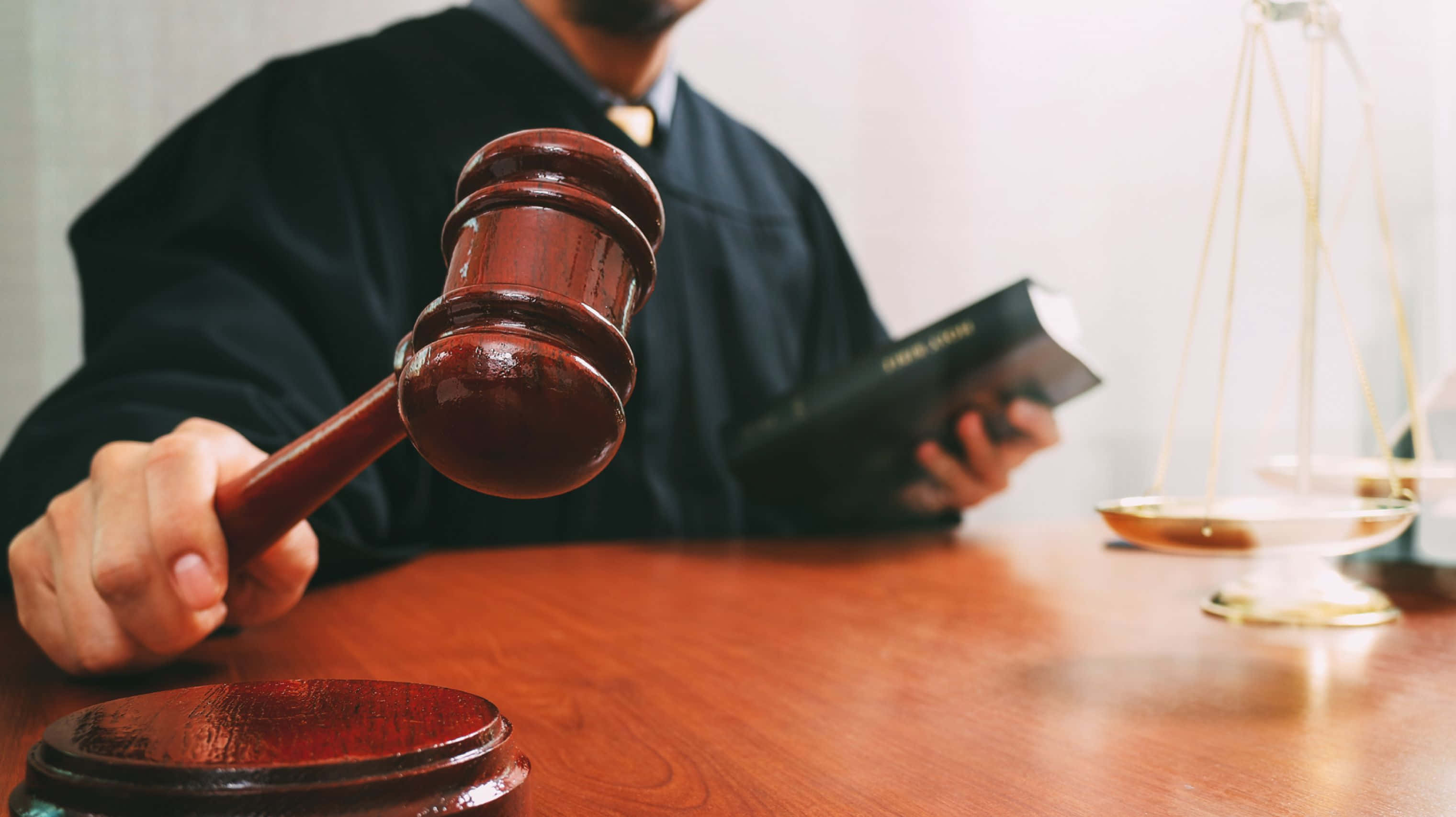 Law 桌布 壁紙 — 一位身穿黑色長袍的法官，手持木槌高舉於音塊之上，同時拿著一本書，背景中有正義的天秤。