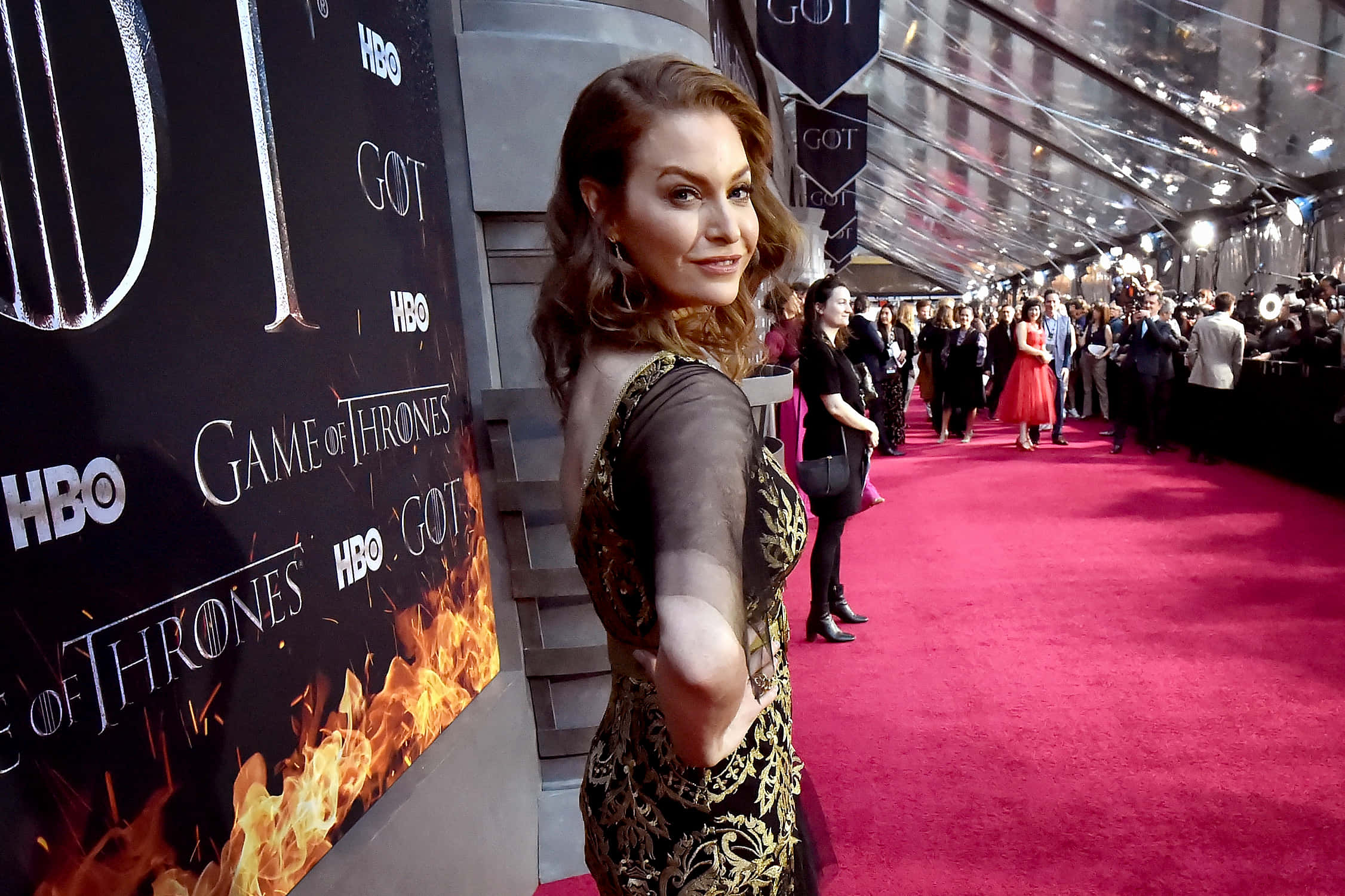Esmé Bianco 桌布 壁紙 — 一位身穿黑金色連衣裙的女性在Game of Thrones HBO活動的鮮豔紅地毯上擺姿勢，背景有火焰和標誌。