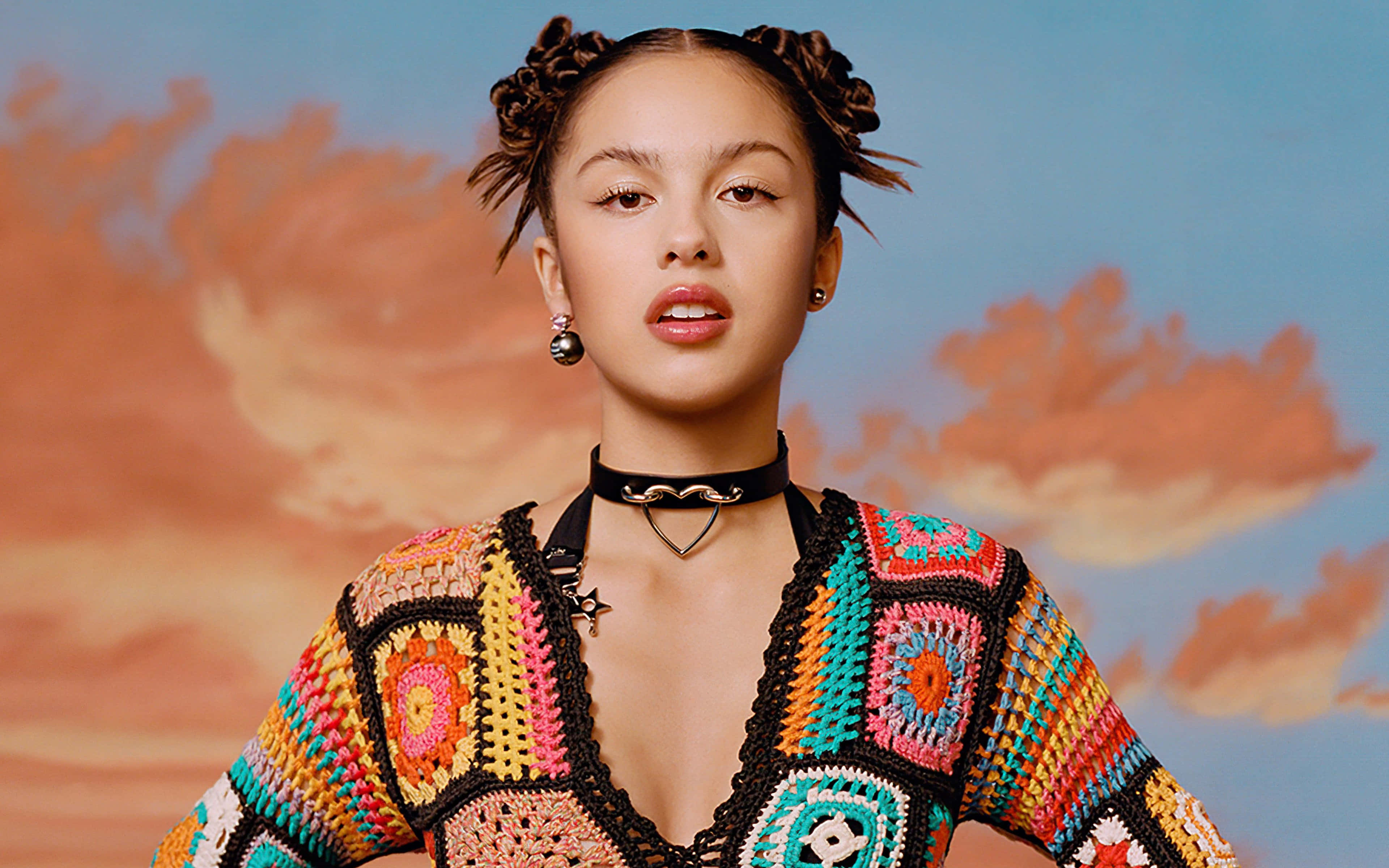 Olivia Rodrigo 桌布 壁紙 — 一位辮子年輕女性穿著色彩繽紛的鉤針上衣，背景是彩繪天空。