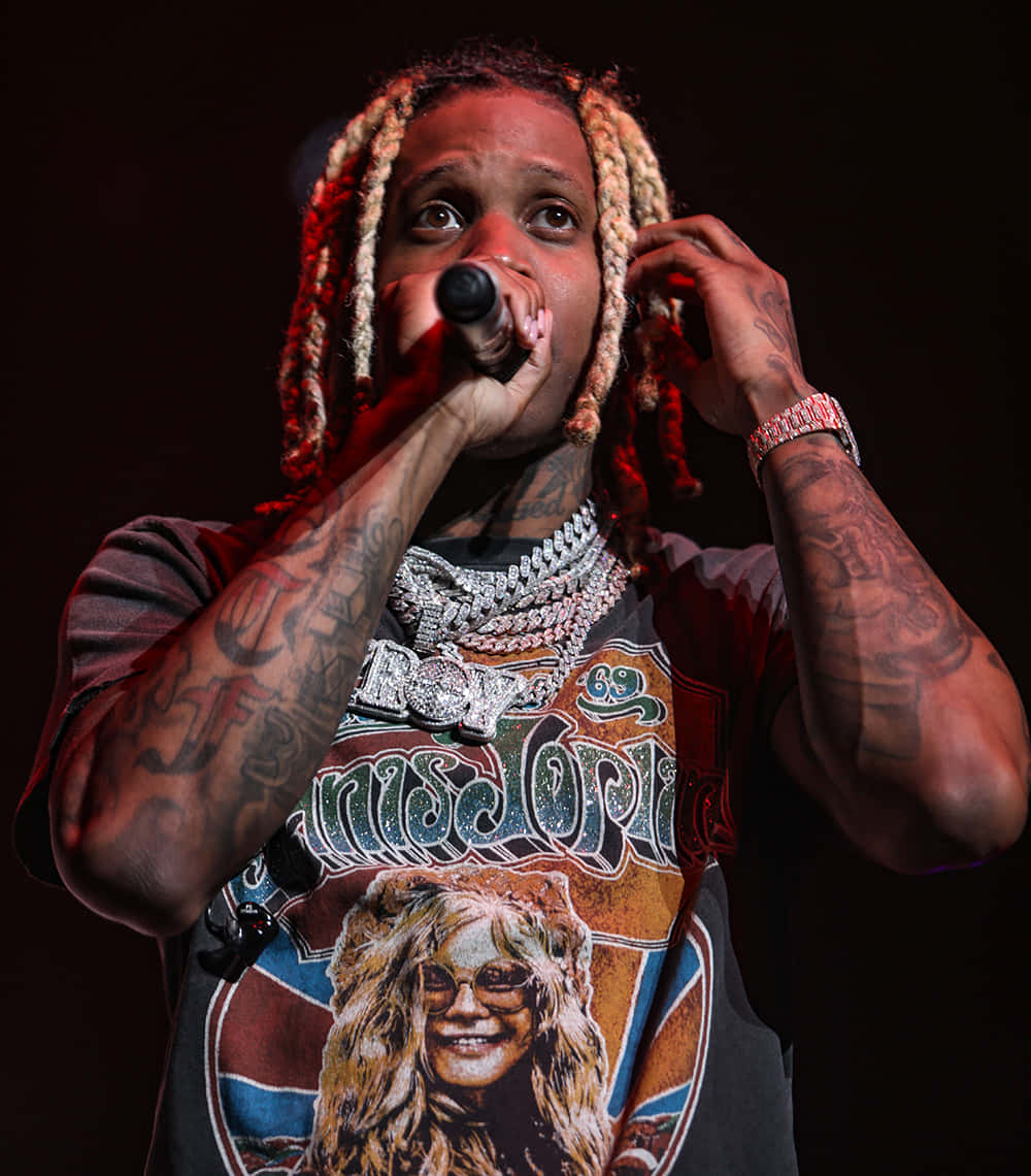 lil durk 桌布 壁紙 — 一位辮子頭髮和紋身的說唱歌手在紅色舞台燈光下拿著麥克風在舞台上表演。