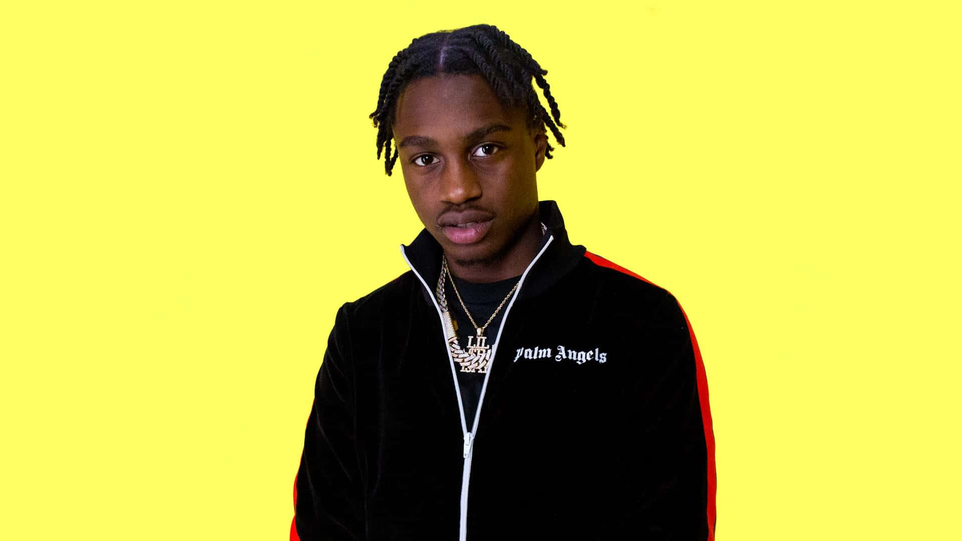 Lil Tjay 桌布 壁紙 — 一位辮子頭髮的年輕男子在明亮的黃色背景前穿著黑色天鵝絨Palm Angels外套。