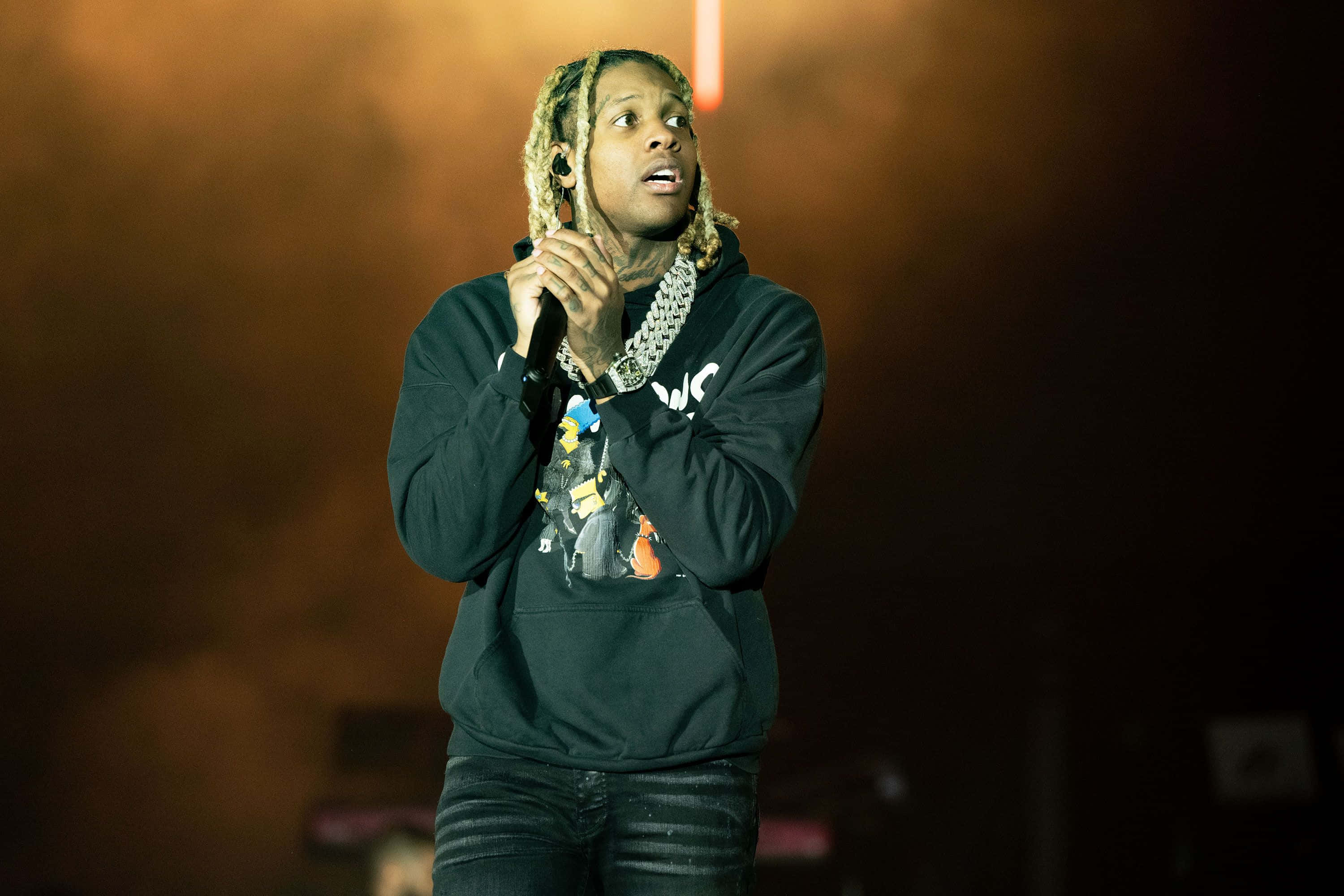 lil durk 桌布 壁紙 — 一位辮子髮型的說唱歌手在溫暖的橙色燈光下拿著麥克風在舞台上表演。