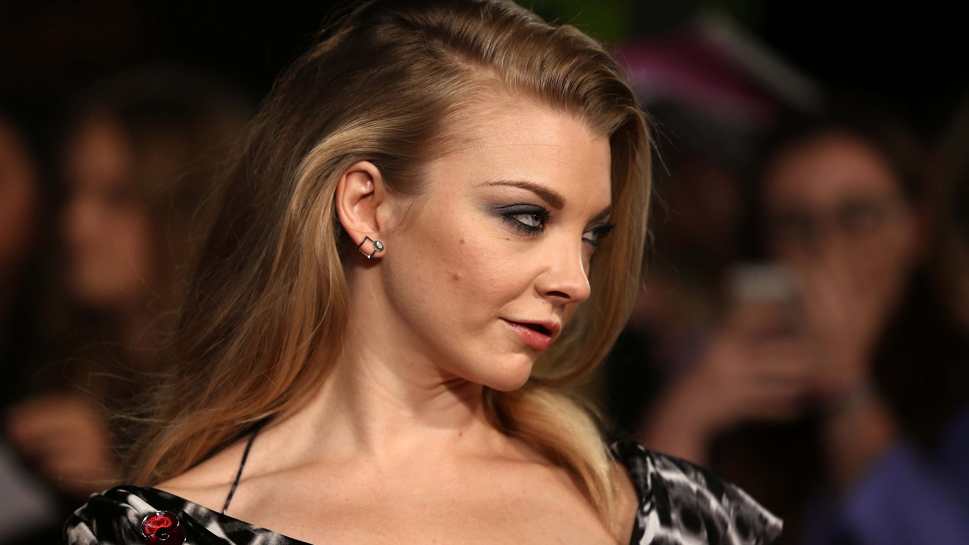 natalie dormer 桌布 壁紙 — 一位迷人嘅金髮女郎，化咗煙燻眼妝，著住黑色圖案連身裙，喺擠迫嘅活動中側視。