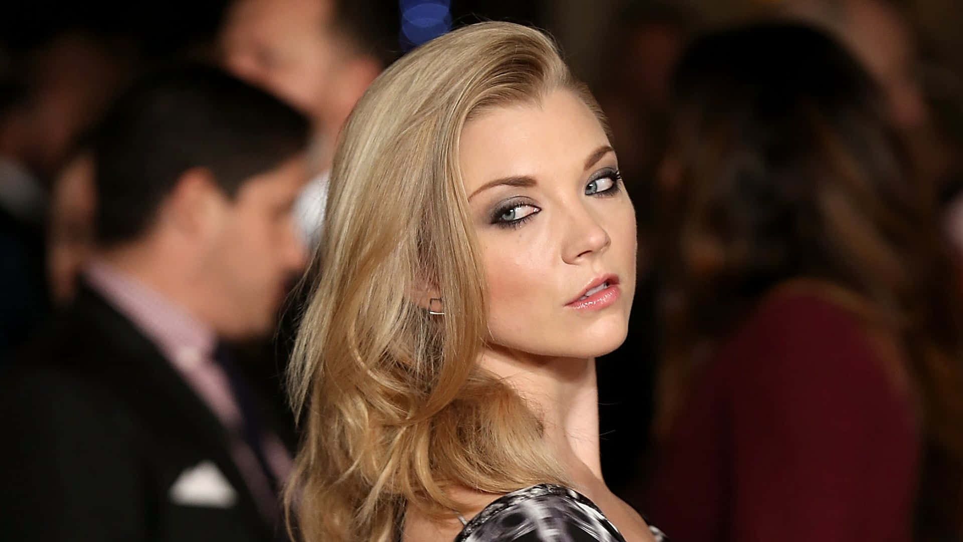 natalie dormer 桌布 壁紙 — 一位迷人金髮女子，煙燻眼妝，在溫暖的環境燈光下，於擁擠的活動中回頭凝視。