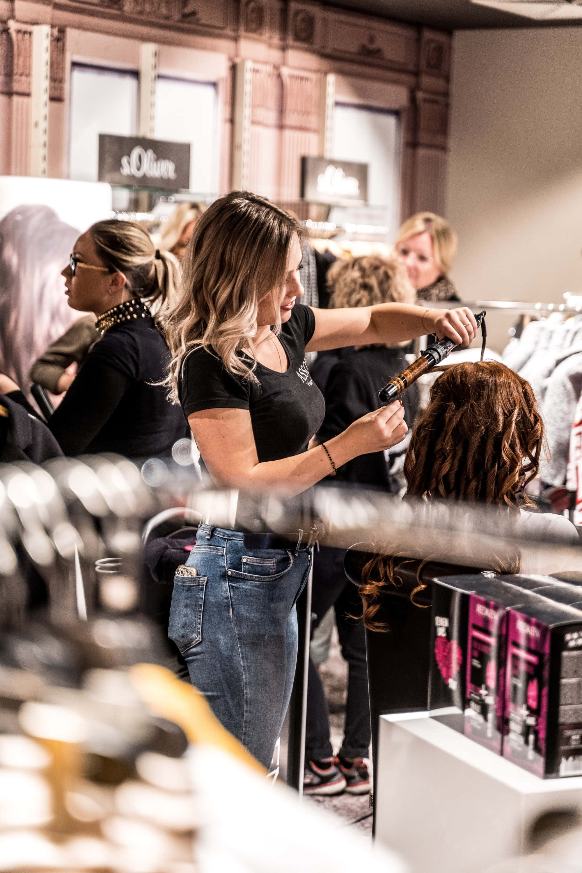 Hair Salon 桌布 壁紙 — 一位造型師在光線充足的精品店中，背景有衣服架和其他顧客，為客戶的頭髮使用捲髮棒。