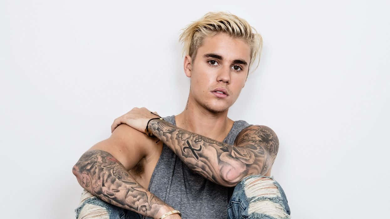 Justin Bieber 桌布 壁紙 — 一位金髮、滿臂紋身的年輕男子，身穿灰色背心和破洞牛仔褲，在白色背景前擺姿勢。