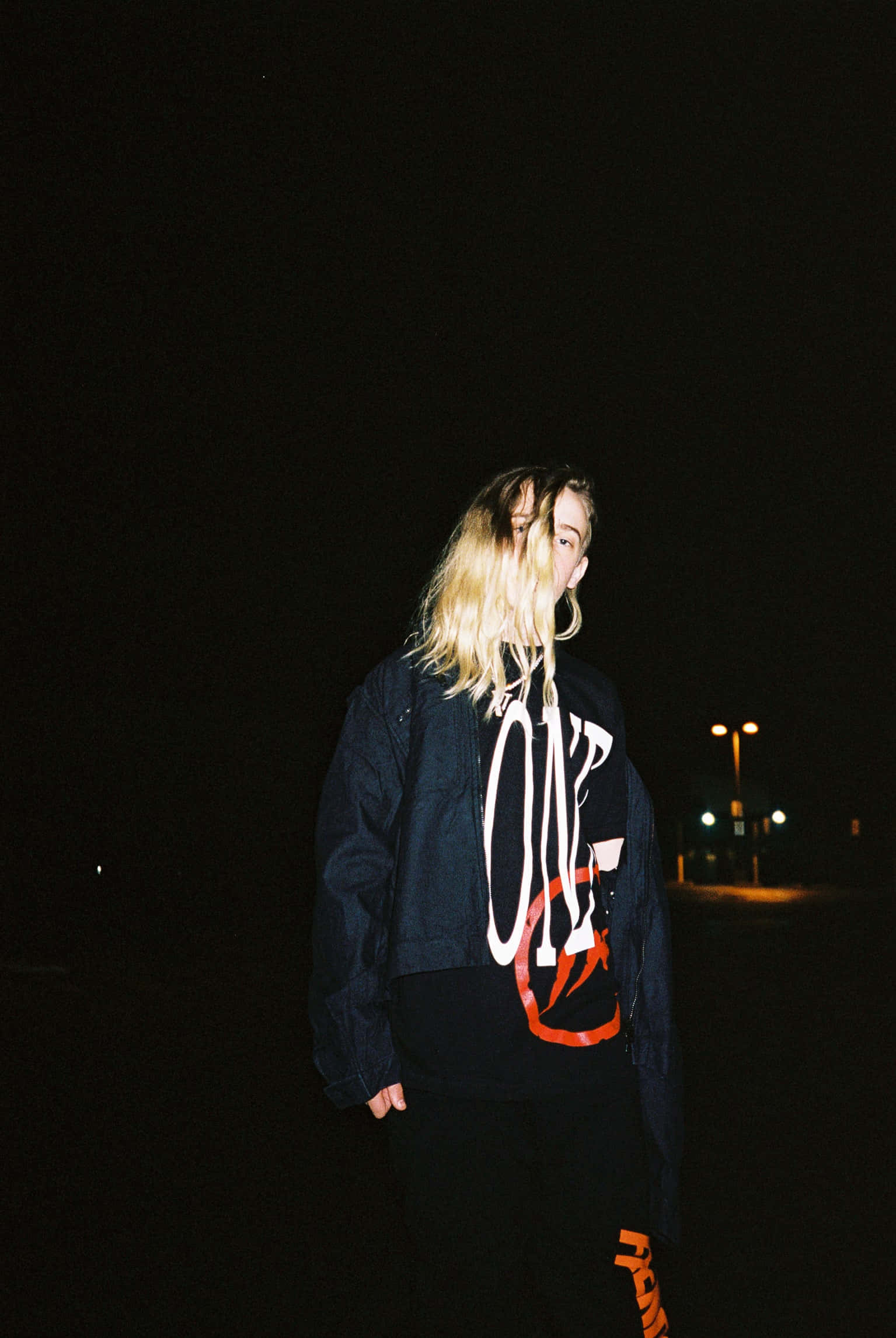 Vlone 桌布 壁紙 — 一位金髮人士站在黑暗的城市夜景中，穿著圖案連帽衫，外搭牛仔外套。