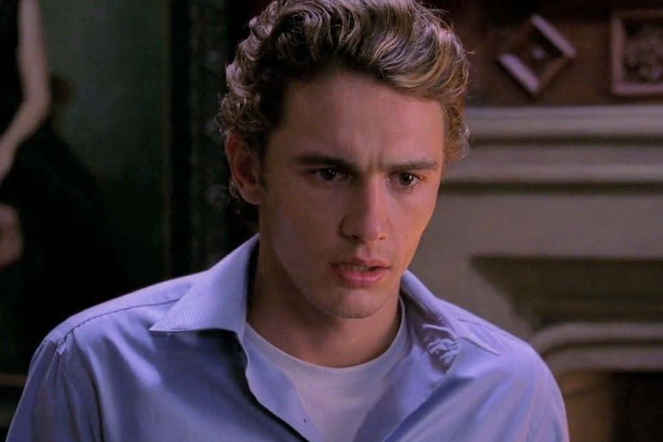 Harry Osborn 桌布 壁紙 — 一位金髮凌亂的年輕男子穿著淺藍色襯衫，在昏暗的房間裡專注地向前凝視。