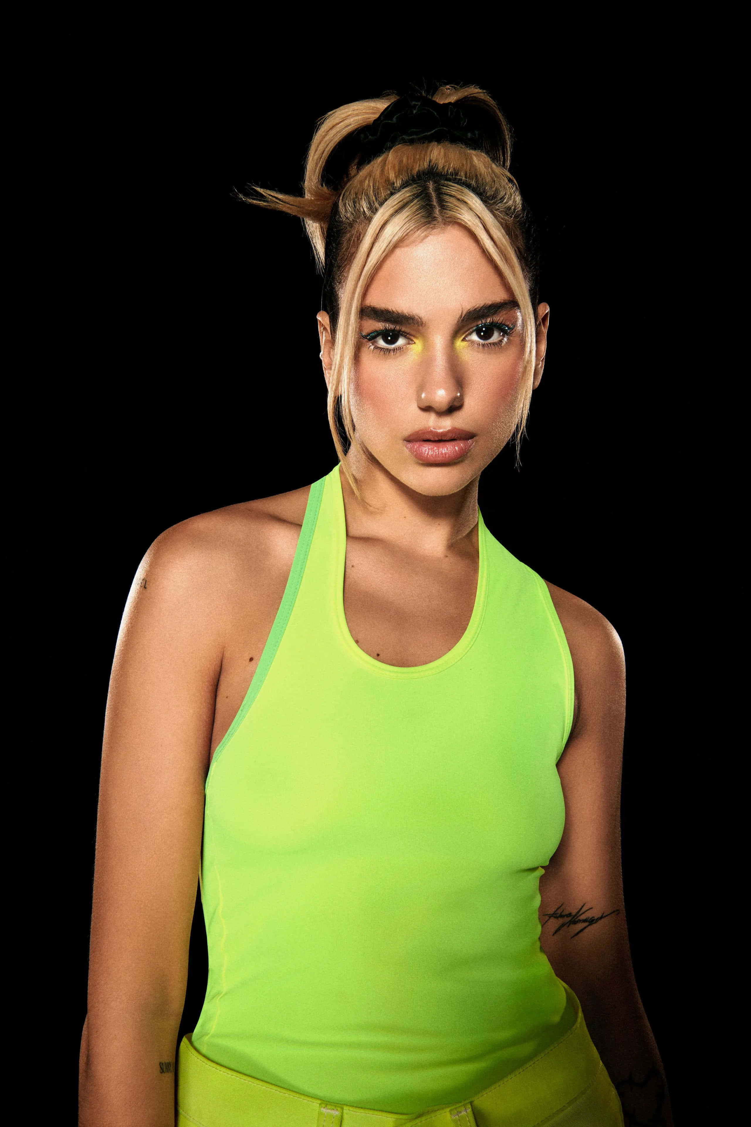 Dua Lipa 桌布 壁紙 — 一位金髮凌亂髮髻的女性，在黑色背景前穿著螢光綠色掛頸上衣。