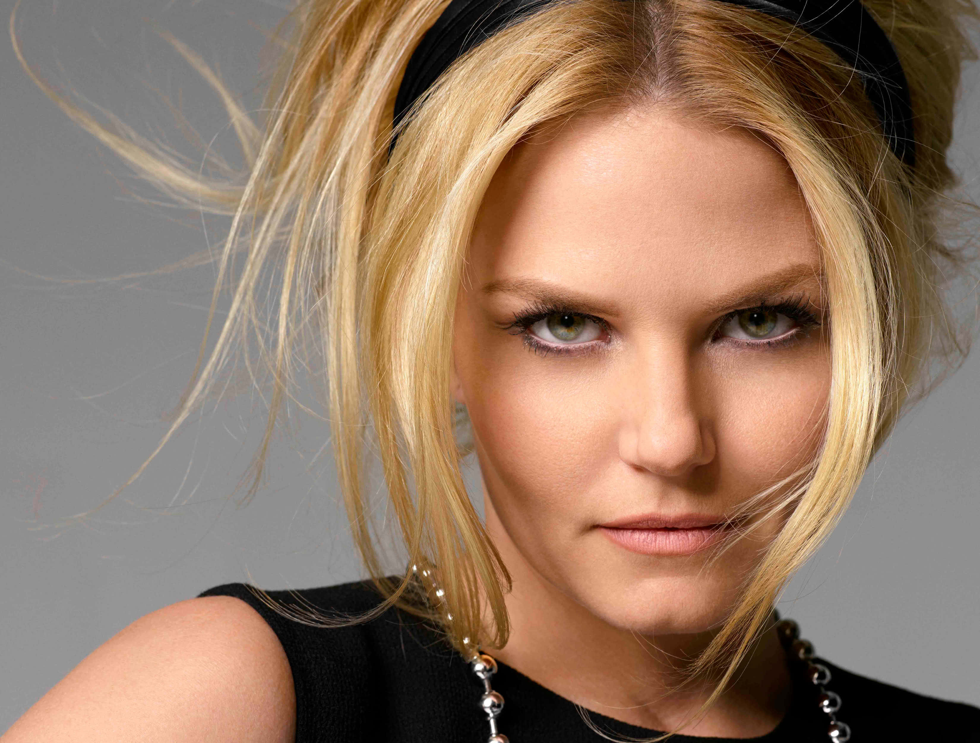 jennifer morrison 桌布 壁紙 — 一位金髮女子，眼神銳利，頭髮被風吹拂，在灰色中性背景前直視鏡頭。