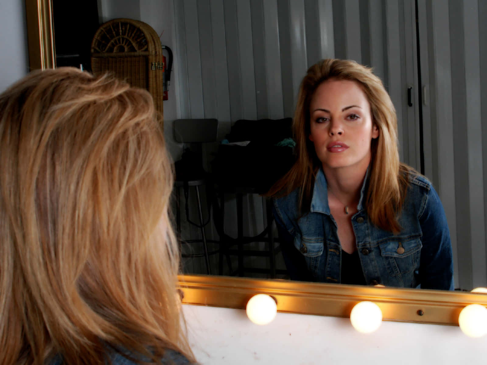 Chandra West 桌布 壁紙 — 一位金髮女子專注地凝視著鑲有發光燈泡的鏡子中的倒影。