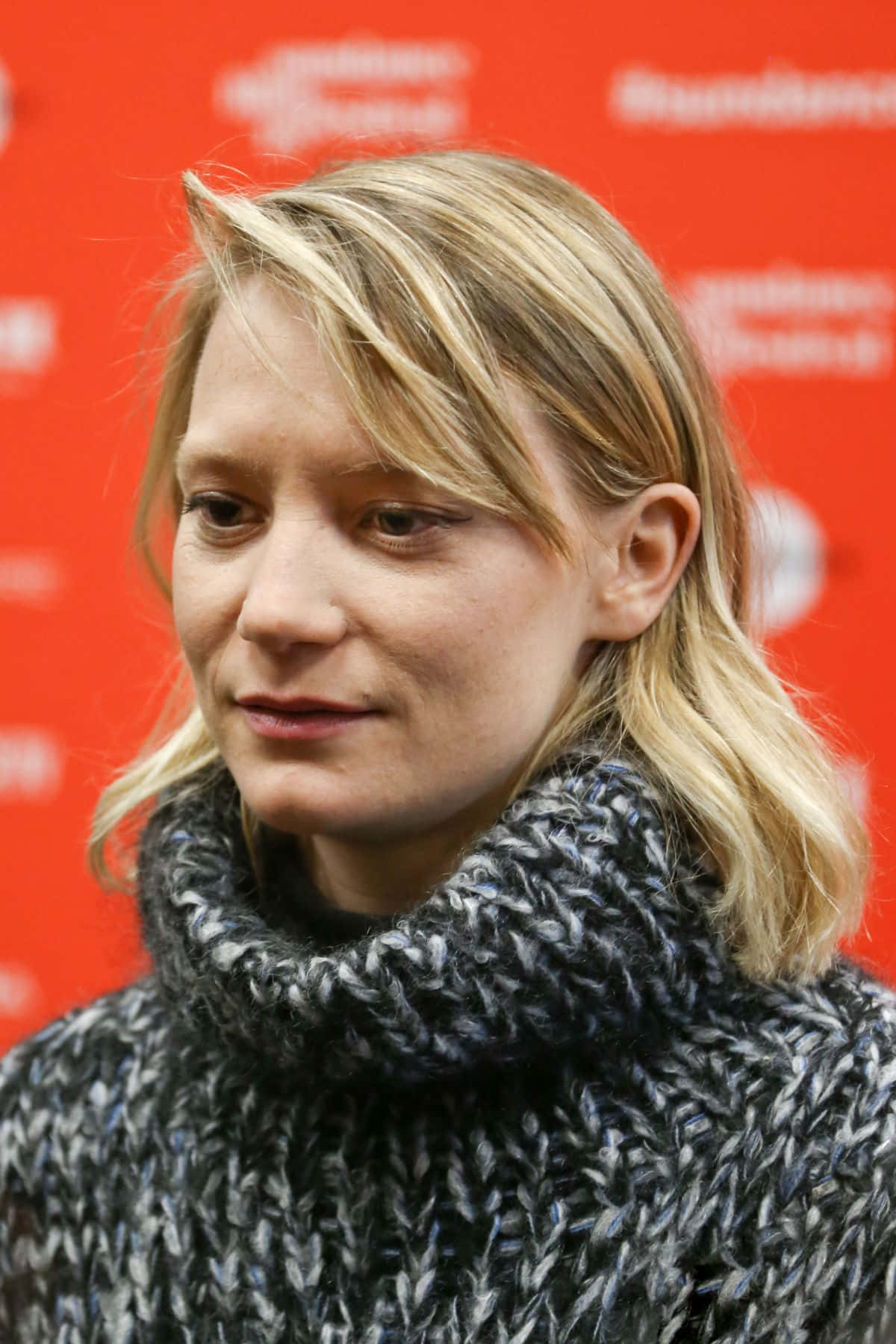 Mia Wasikowska 桌布 壁紙 — 一位金髮女子戴著厚織圍巾，在鮮紅色的品牌背景前若有所思地凝視。
