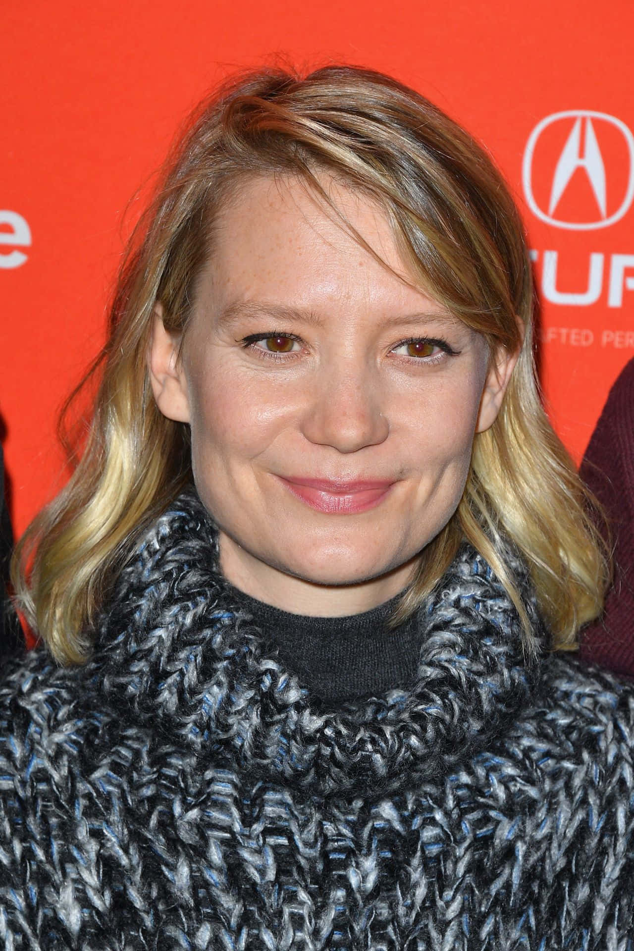 Mia Wasikowska 桌布 壁紙 — 一位金髮女子戴著針織圍巾，在鮮紅色品牌背景前溫柔微笑。
