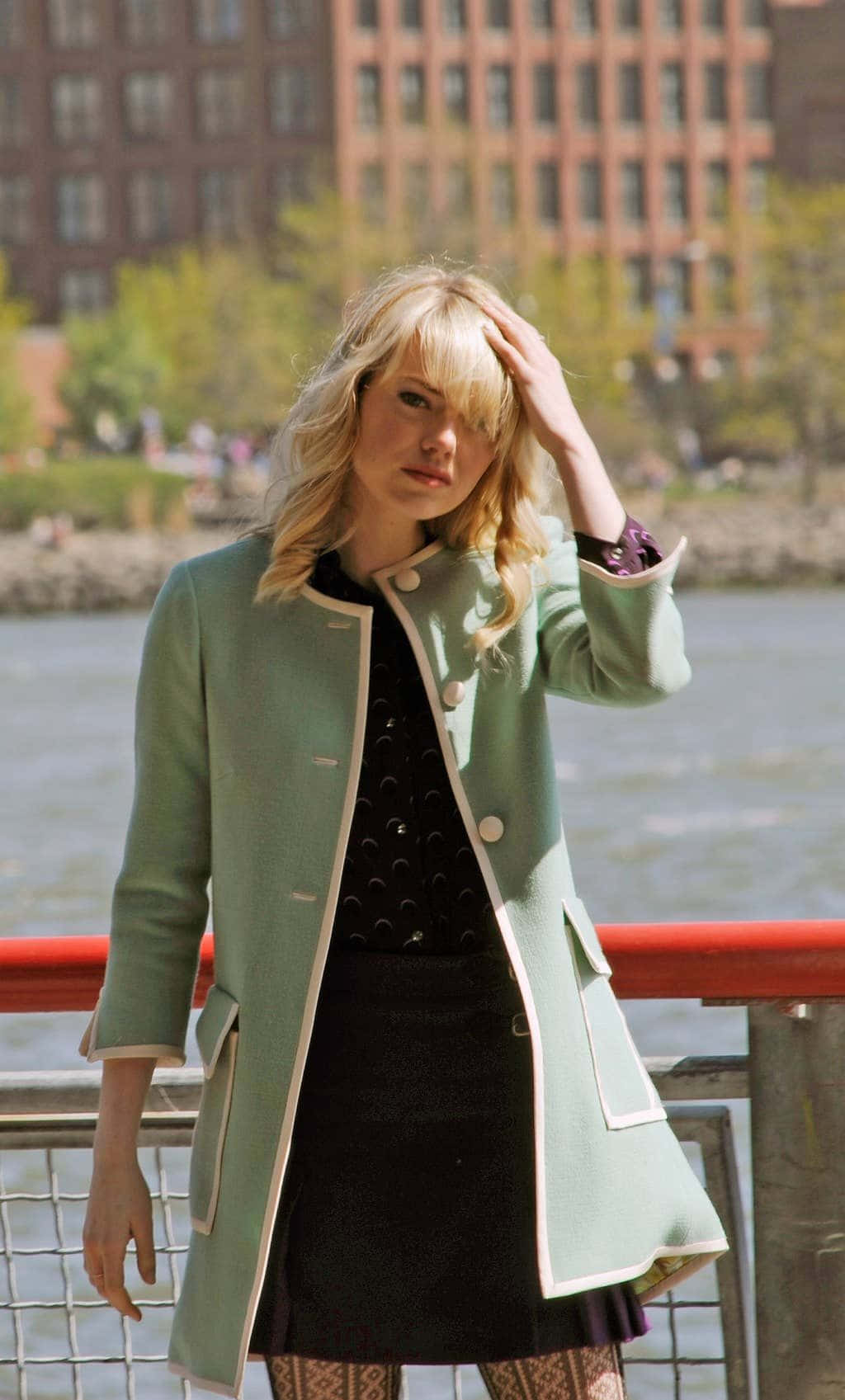 Gwen Stacy 桌布 壁紙 — 一位金髮女子穿著薄荷綠色外套，在陽光明媚的日子裡站在河邊欄杆旁，身後是城市建築。