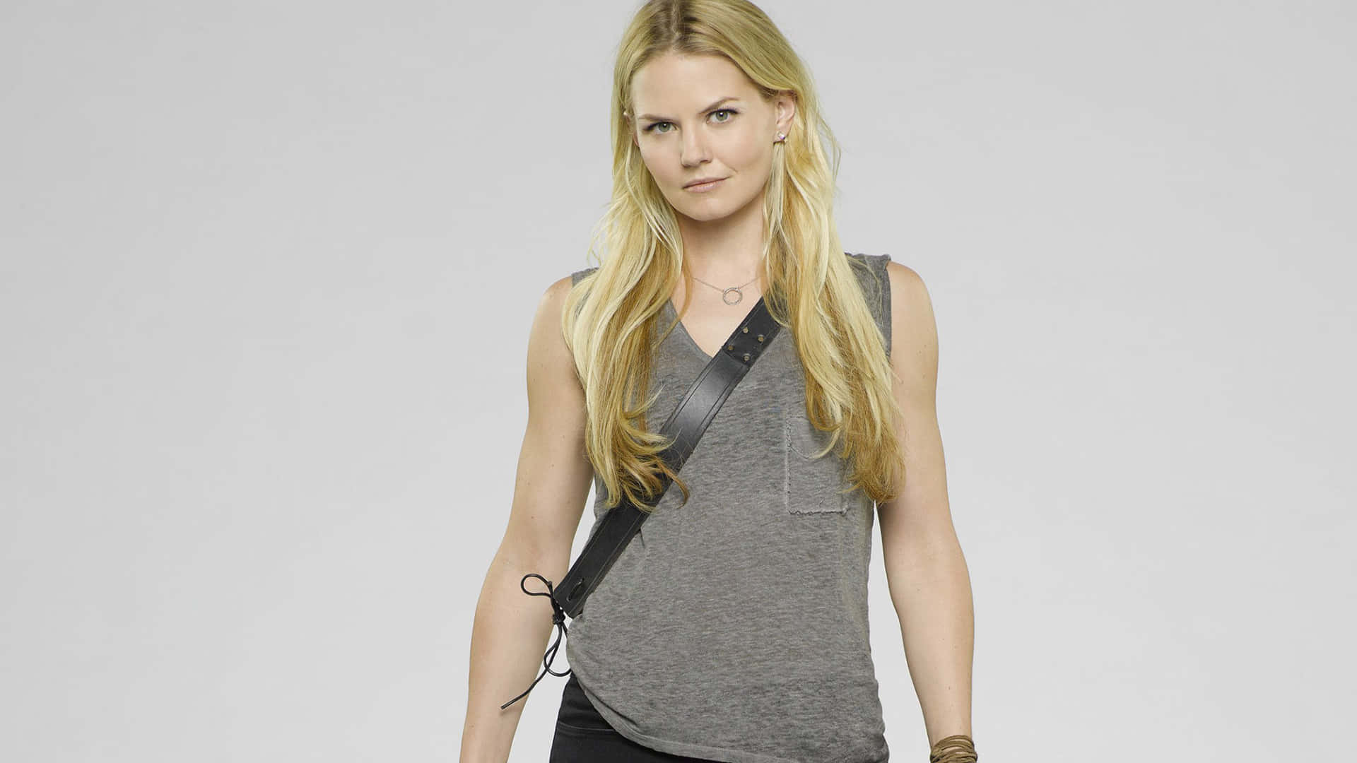 jennifer morrison 桌布 壁紙 — 一位金髮女子表情嚴肅，身穿灰色背心，背著黑色斜挎包，背景是純白色。