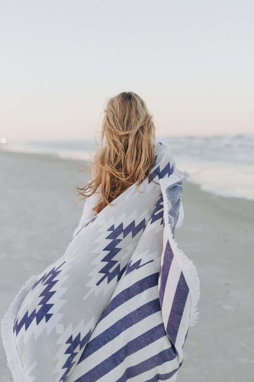 Winter Beach 桌布 壁紙 — 一位金髮女子裹著藍白圖案毛毯，在日落時分站在沙灘上面向海洋。
