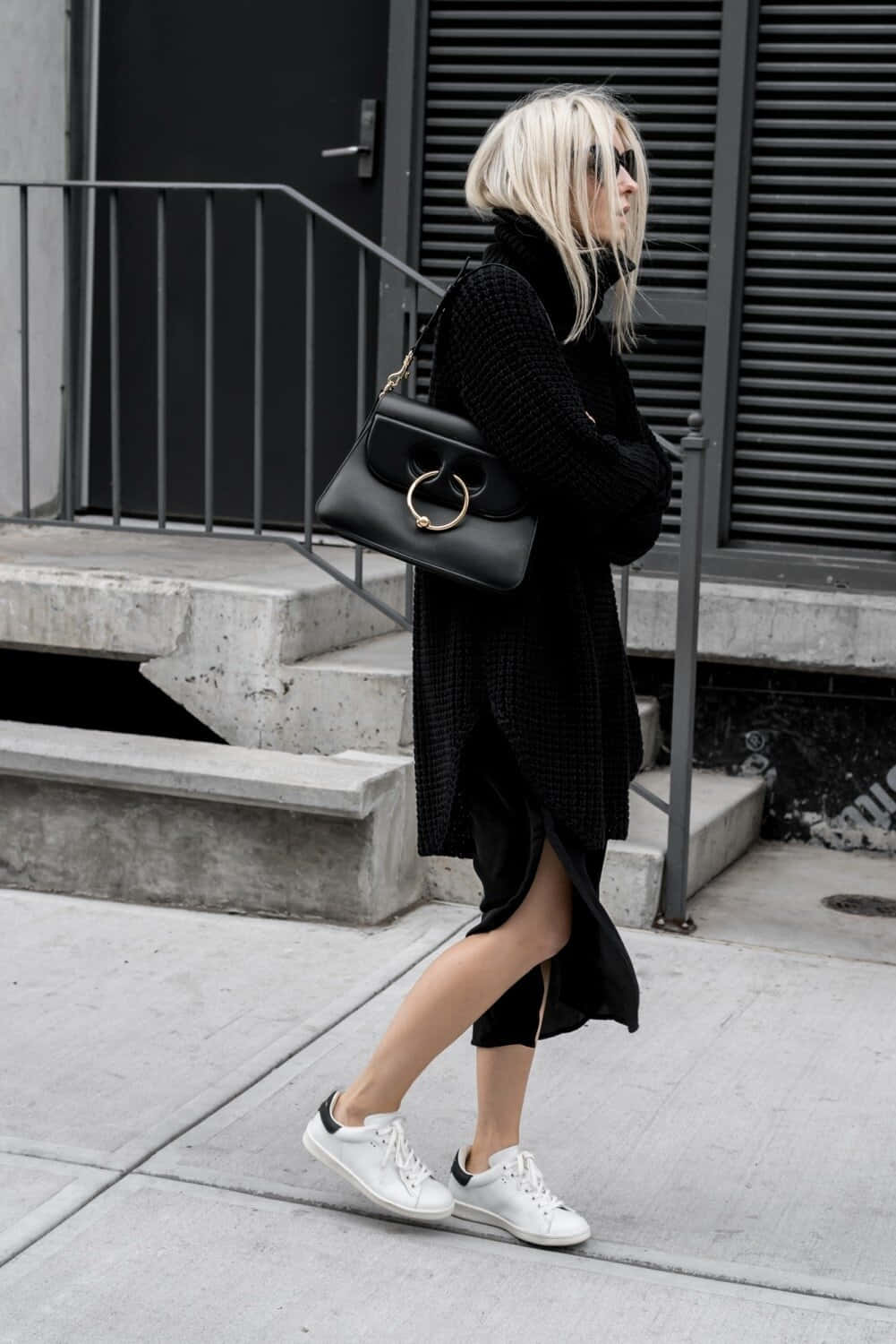 Minimalist Fashion 桌布 壁紙 — 一位金髮女子身穿全黑衣服，走過混凝土行人路，經過金屬樓梯和深色建築物外牆。