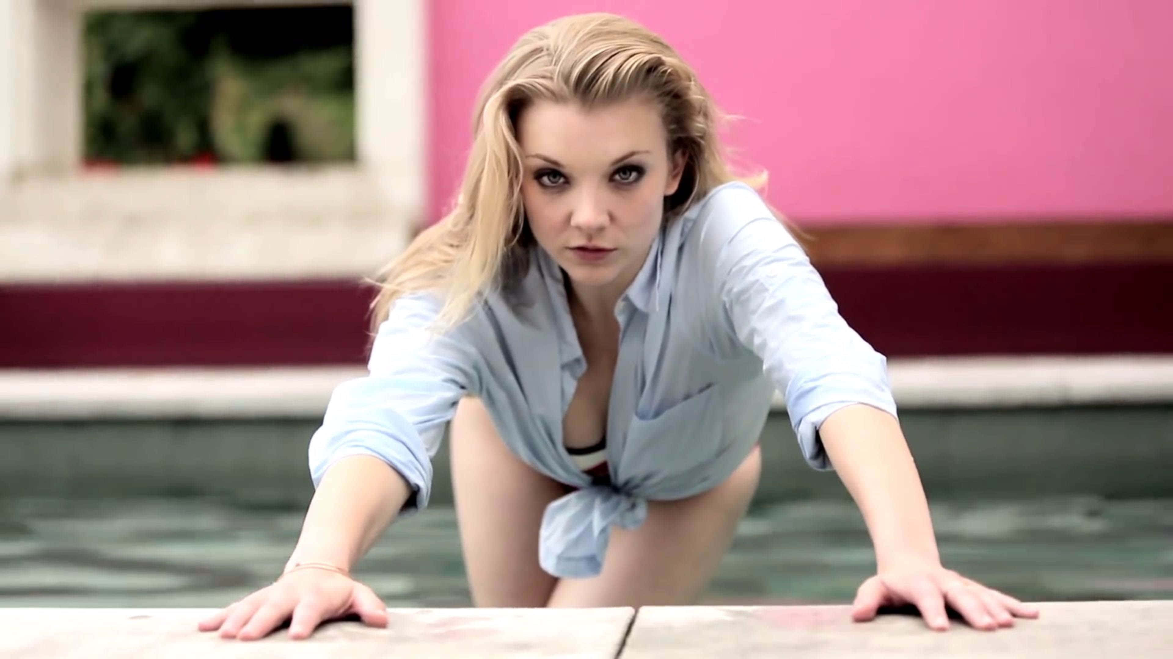 natalie dormer 桌布 壁紙 — 一位金髮女子身穿淺藍色襯衫，在粉紅色牆壁的背景下，身體前傾靠在泳池邊緣。
