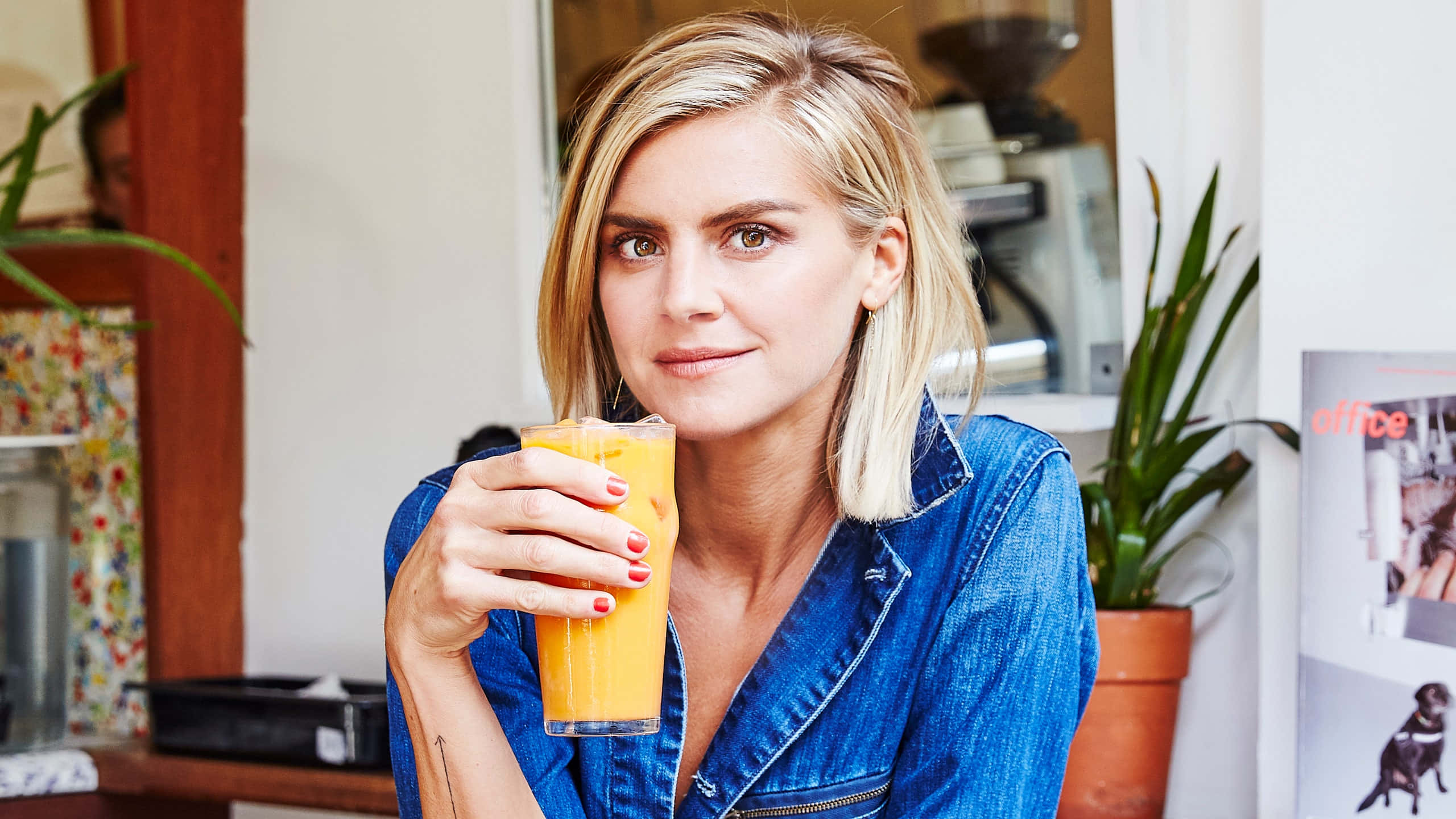 eliza coupe 桌布 壁紙 — 一位金髮女子身穿牛仔外套，在舒適的咖啡廳環境中手持一杯橙汁，對著鏡頭微笑。