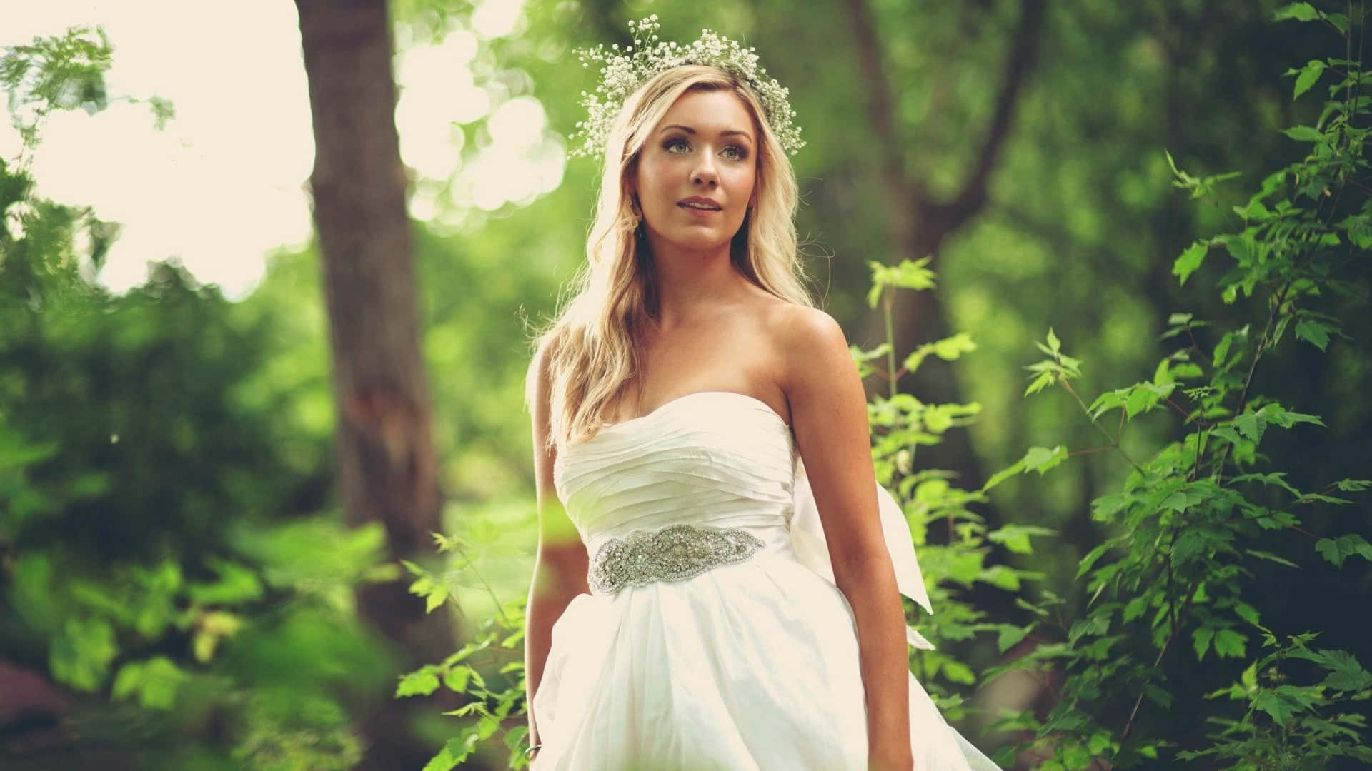 Wedding Dress 桌布 壁紙 — 一位金髮女子身穿白色無肩帶婚紗，頭戴花冠，優雅地站在陽光灑落的森林中。