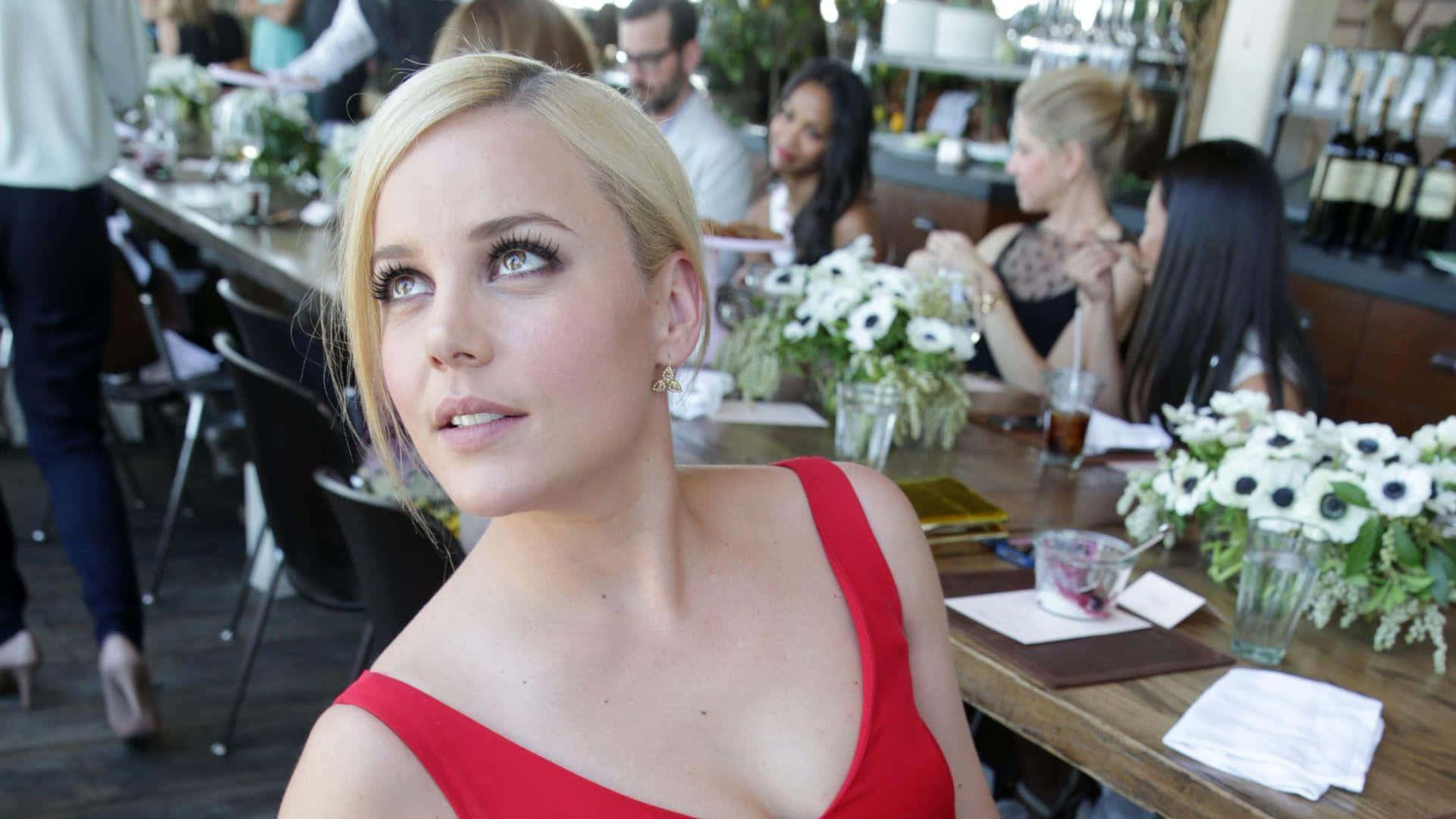 Abbie Cornish 桌布 壁紙 — 一位金髮女子身穿紅色連衣裙，在一個熙熙攘攘的戶外餐廳中向上凝視，背景有白色花朵和顧客。