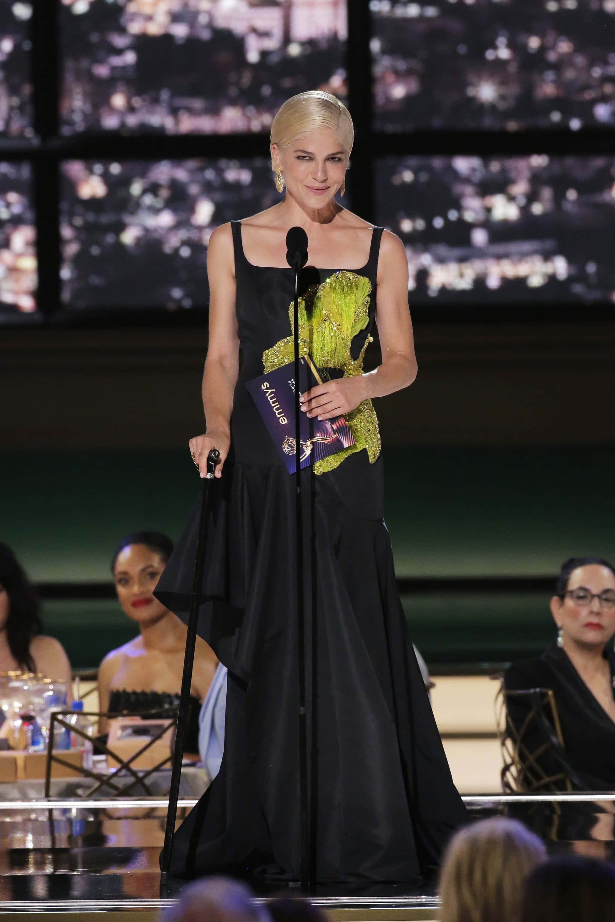 Selma Blair 桌布 壁紙 — 一位金髮女子身穿黑色長袍，上面有綠色閃片龍形圖案，站在講台上，在都市天際線背景的舞台上手持獎項。
