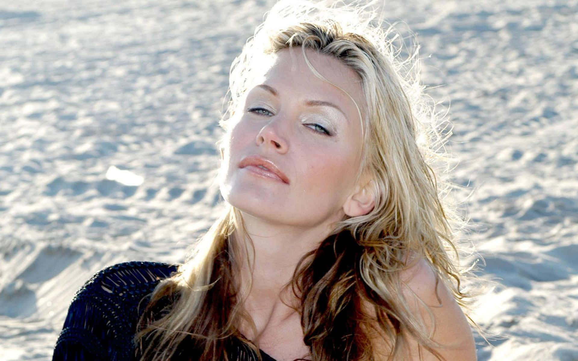 Natasha Henstridge 桌布 壁紙 — 一位金髮女子閉著眼睛在沙灘上享受陽光，身穿深色質感上衣。