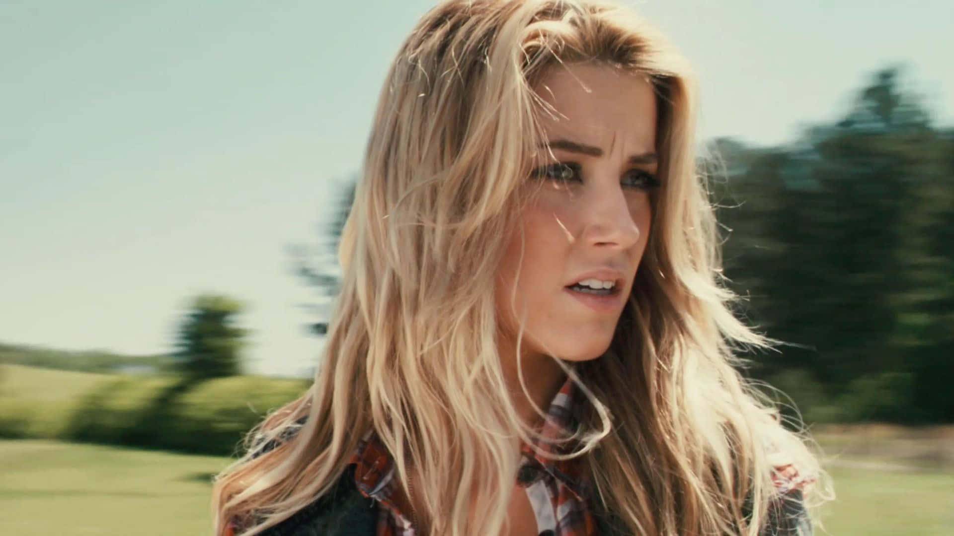 Amber Heard 桌布 壁紙 — 一位金髮女子頭髮被風吹亂，在陽光明媚的綠色田野中快速移動，表情驚慌。