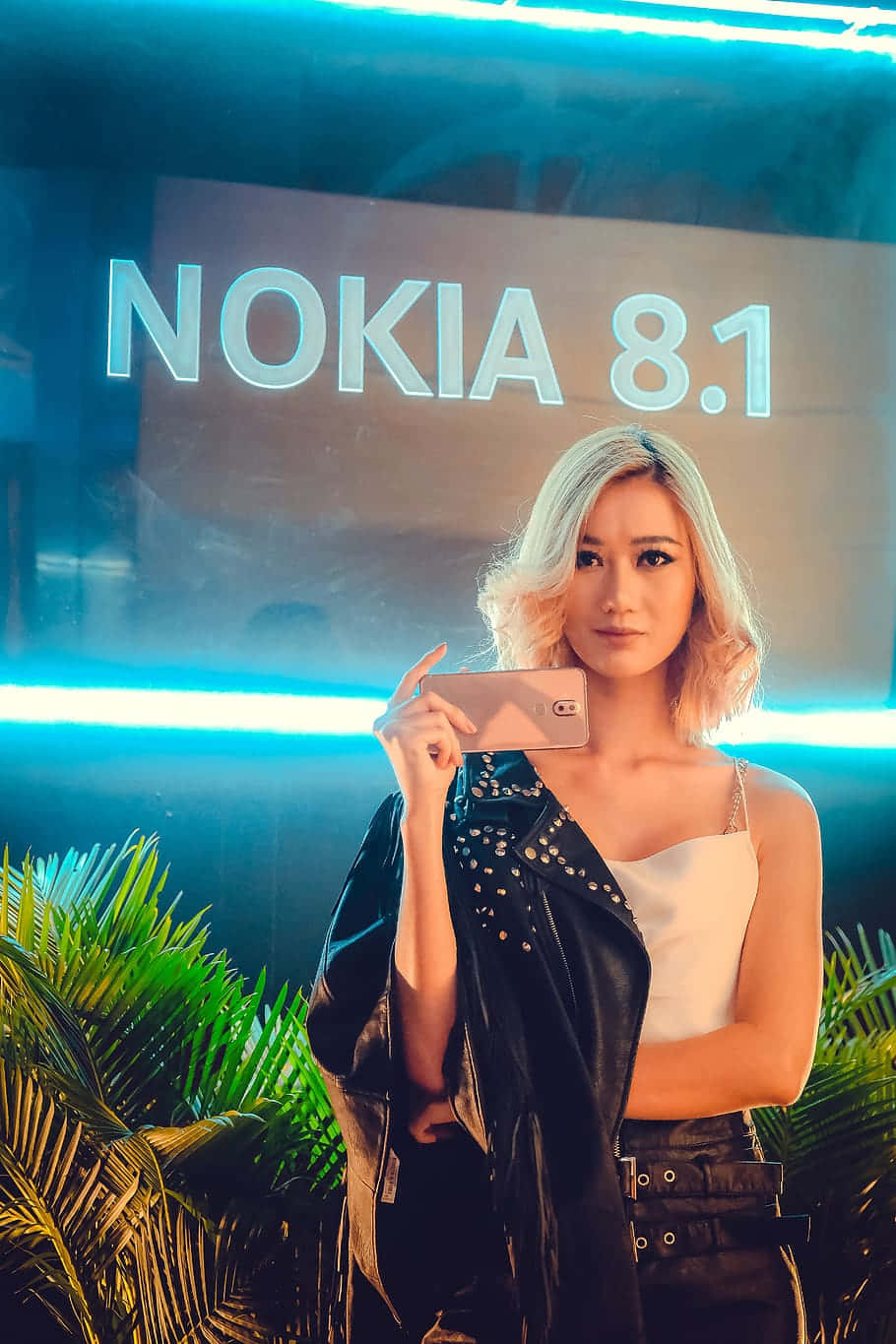 Nokia 桌布 壁紙 — 一位金髮女性在霓虹燈下，於發光的Nokia 8.1標誌前手持電話，周圍環繞著熱帶植物。