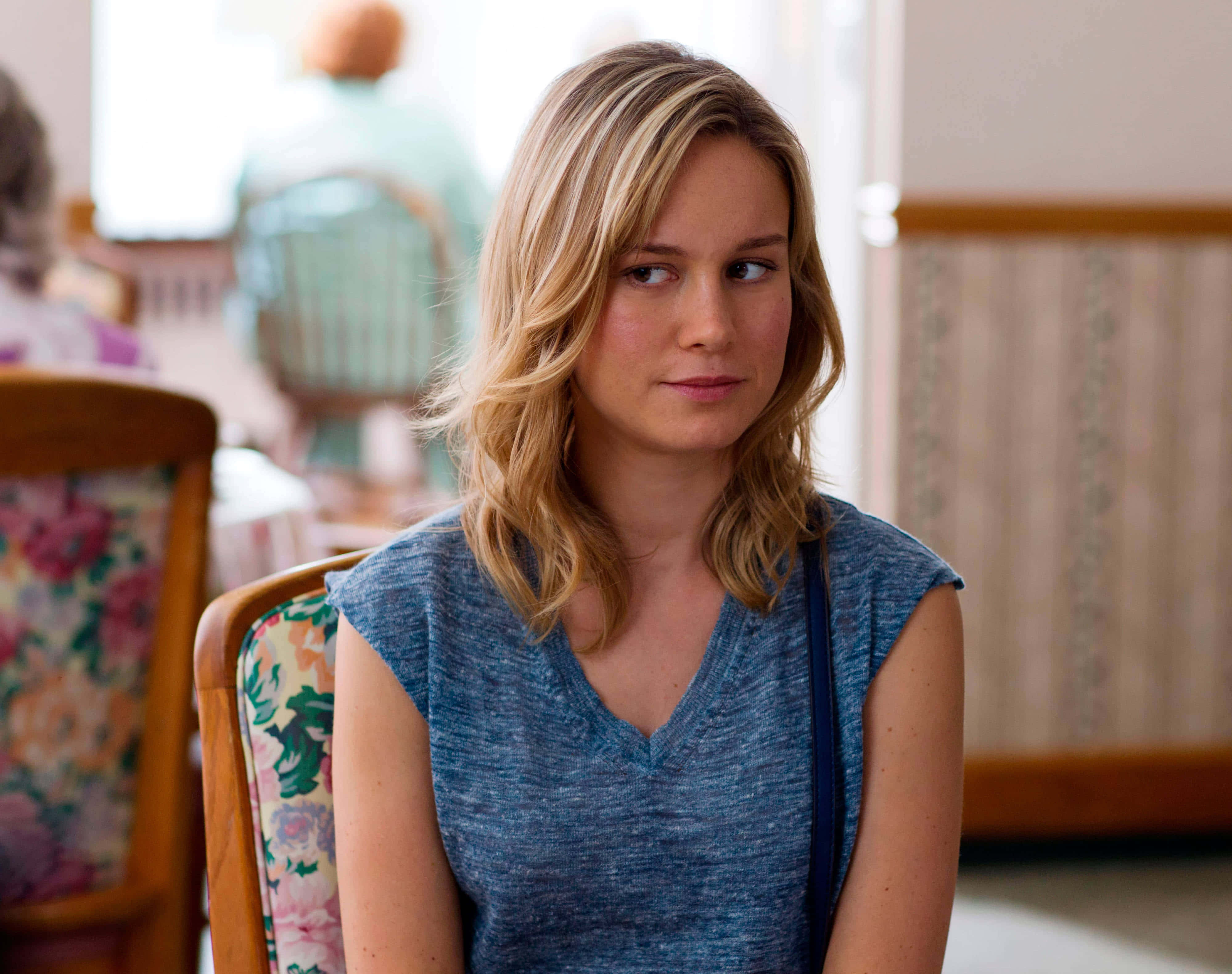 brie larson 桌布 壁紙 — 一位金髮女性坐在花卉圖案的椅子上，在柔和光線的室內環境中看向一旁。