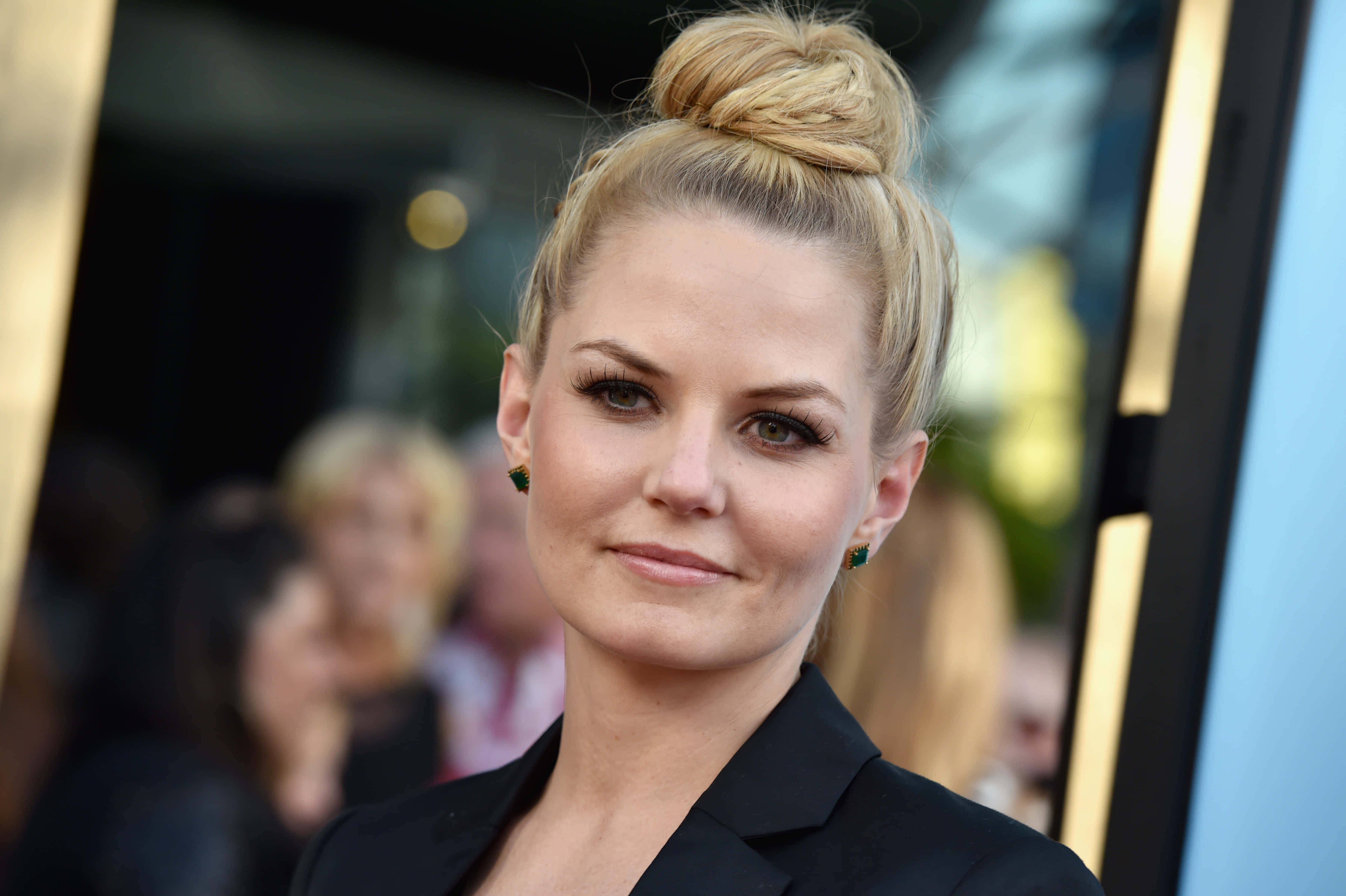 jennifer morrison 桌布 壁紙 — 一位金髮女性將頭髮盤成高髻，穿著黑色西裝外套和綠色耳環，溫柔地微笑。