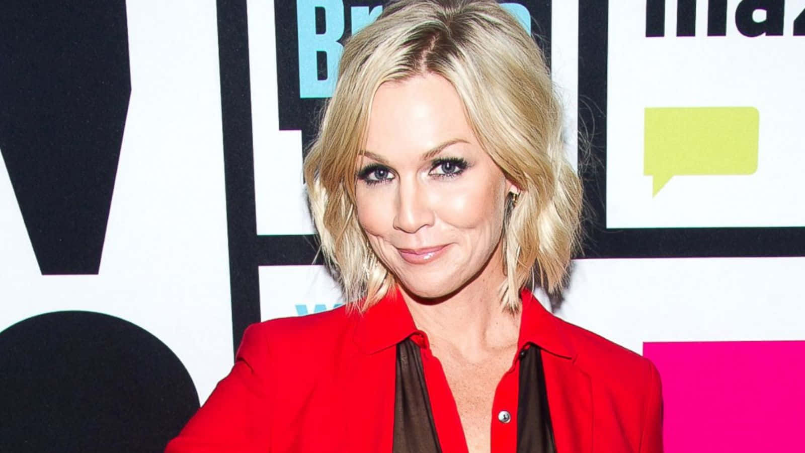 Jennie Garth 桌布 壁紙 — 一位金髮女性穿著大膽的紅色西裝外套，在色彩繽紛的圖形背景前自信地微笑。