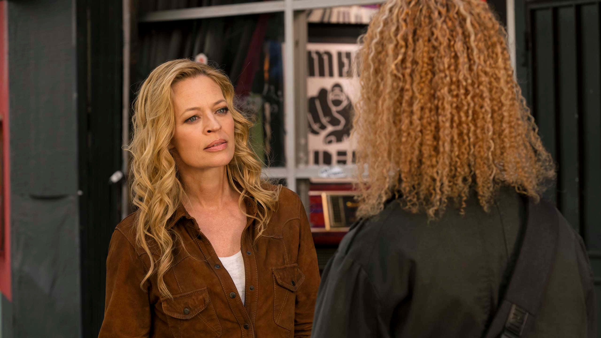 Jeri Ryan 桌布 壁紙 — 一位金髮女性穿著棕色麂皮外套，從後面專注地看著另一個捲髮的人。