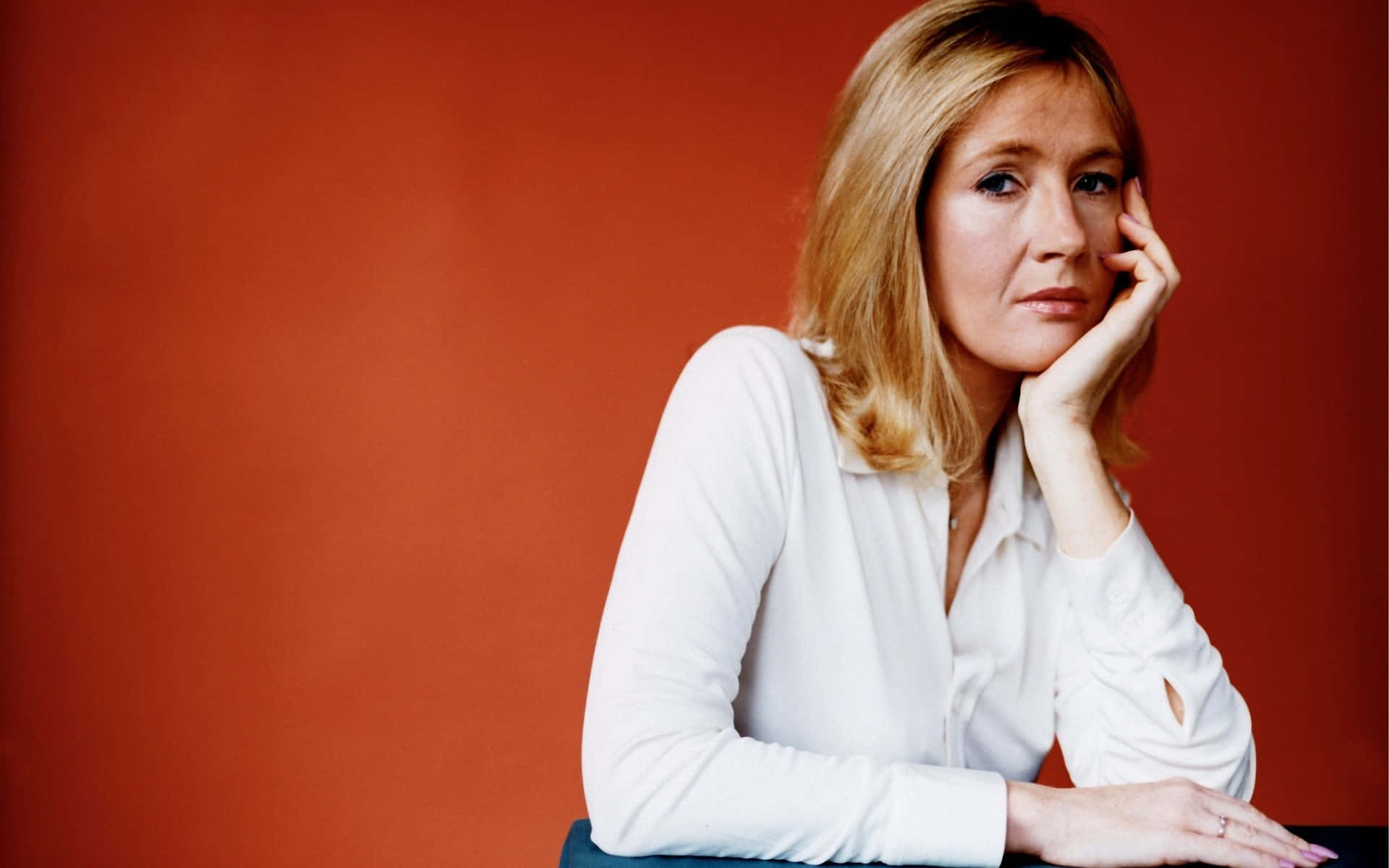J K Rowling 桌布 壁紙 — 一位金髮女性穿著白色襯衫，將下巴靠在手上，倚著一面純橙色牆壁。