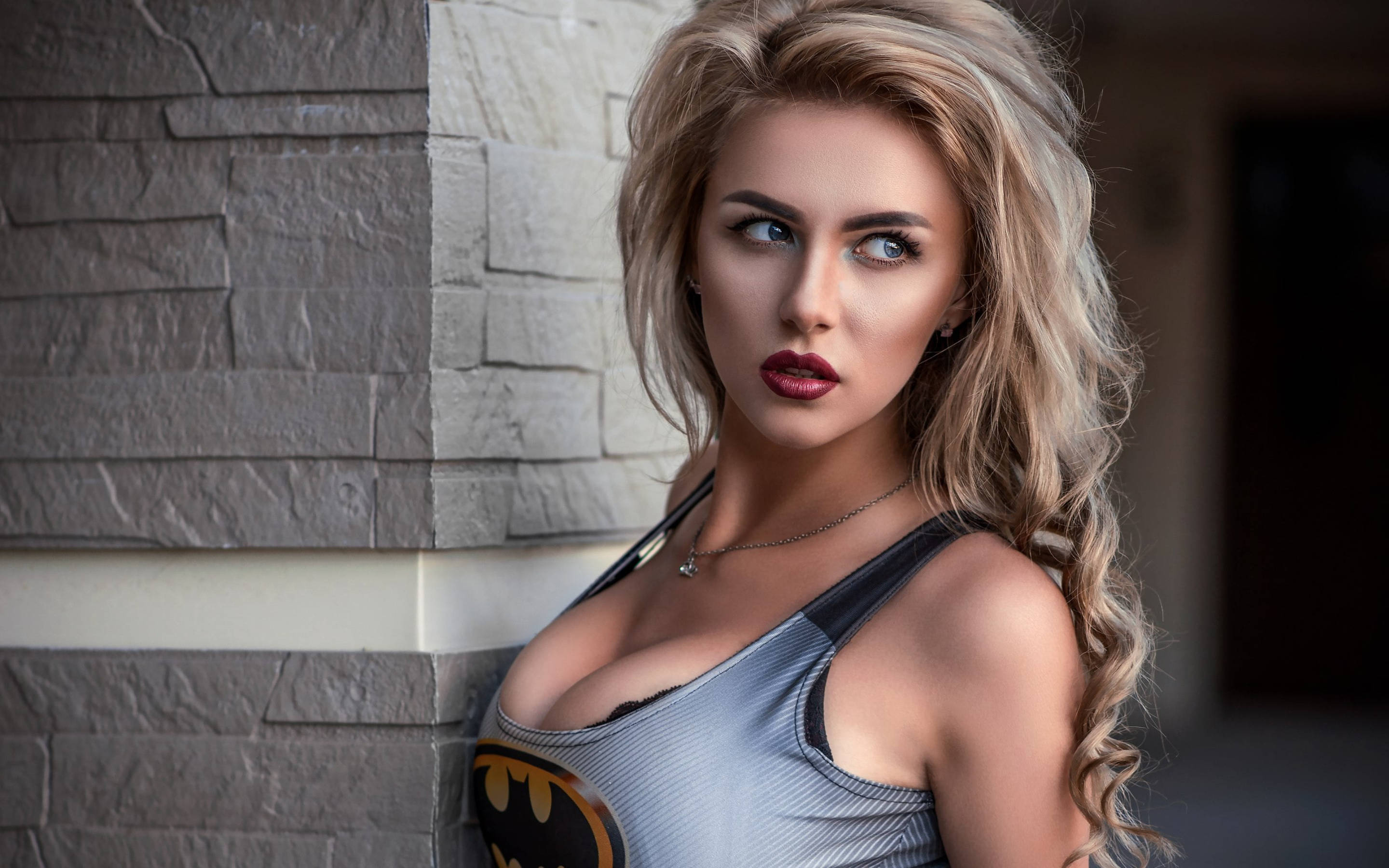 Batman Logo 桌布 壁紙 — 一位金髮女性穿著蝙蝠俠主題上衣，以銳利的眼神靠著石牆。