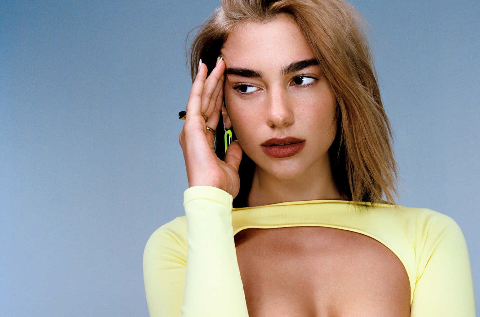 Dua Lipa 桌布 壁紙 — 一位金髮女性穿著黃色上衣，在純藍色背景前將手機貼在耳邊。