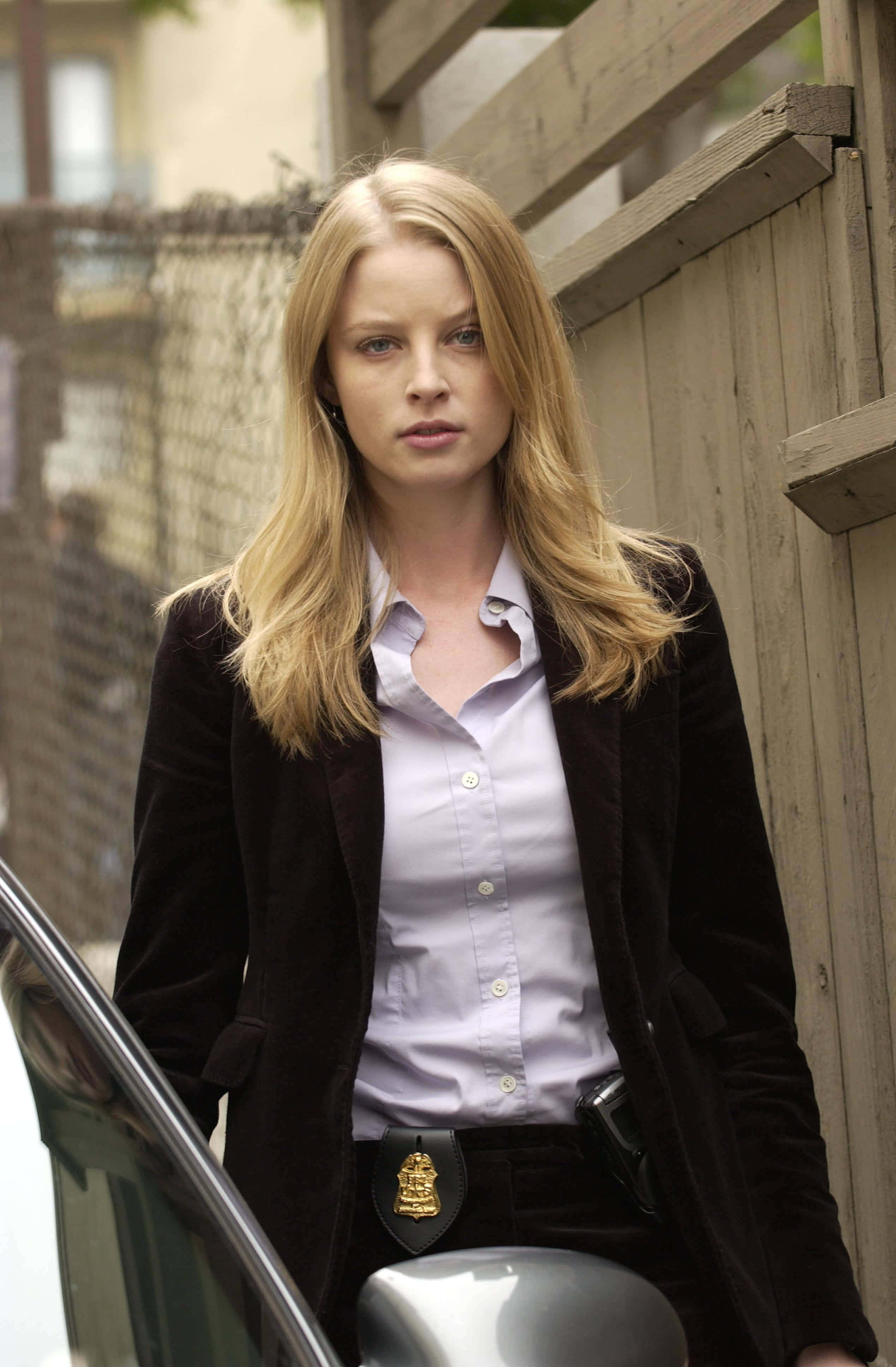 rachel nichols 桌布 壁紙 — 一位金髮女警穿著深色天鵝絨外套，腰帶上可見徽章，站在一輛車旁。