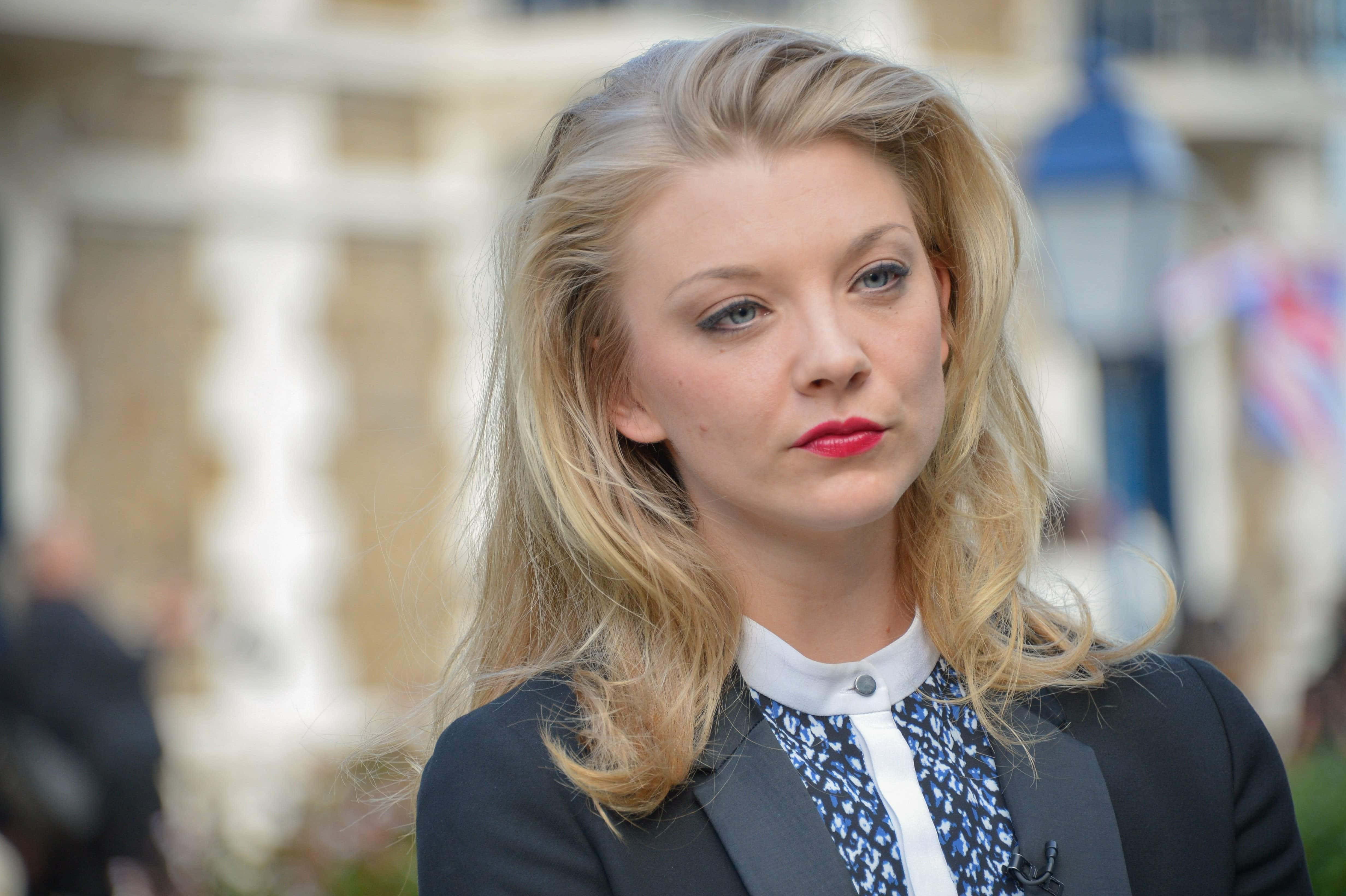 natalie dormer 桌布 壁紙 — 一位金髮女郎，紅唇藍眼，穿著深色西裝外套和圖案襯衫，在戶外若有所思地凝視。
