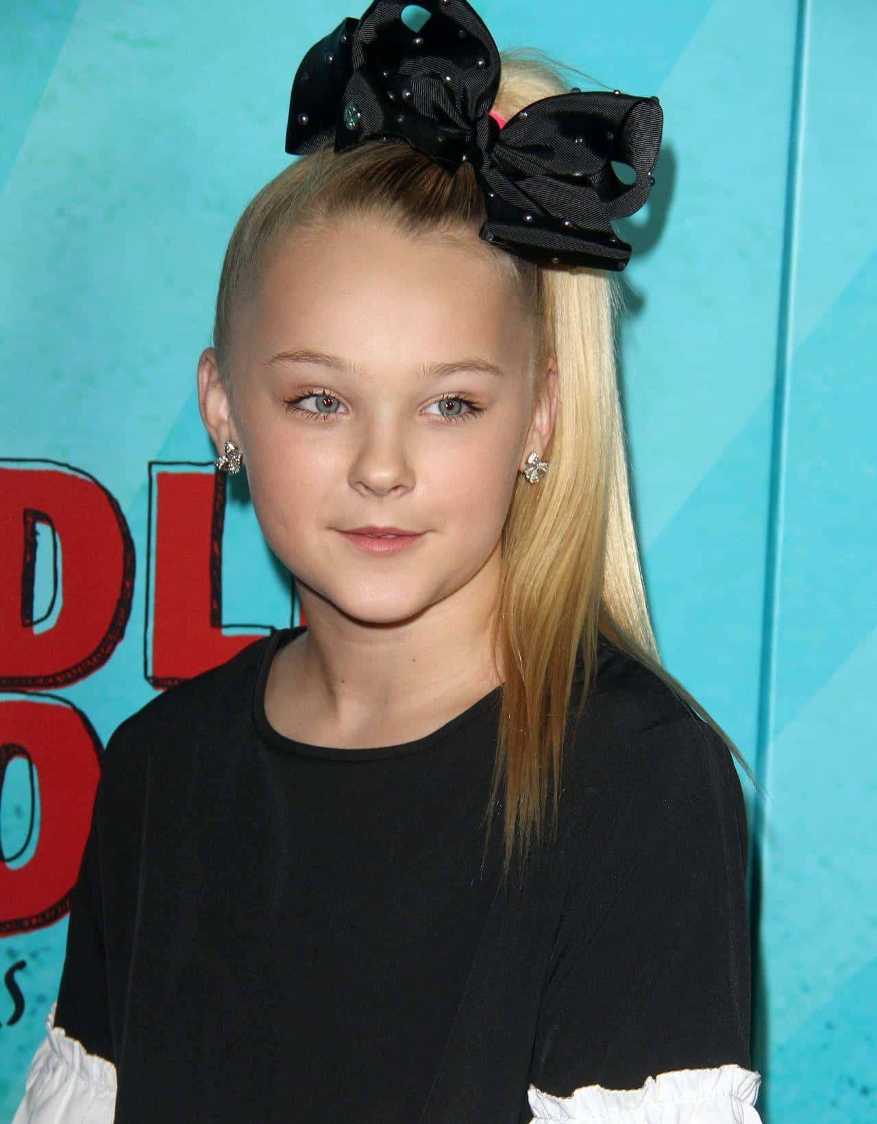 jojo siwa 桌布 壁紙 — 一位金髮少女戴著一個大黑色蝴蝶結，在明亮的藍色背景下對著鏡頭微笑。