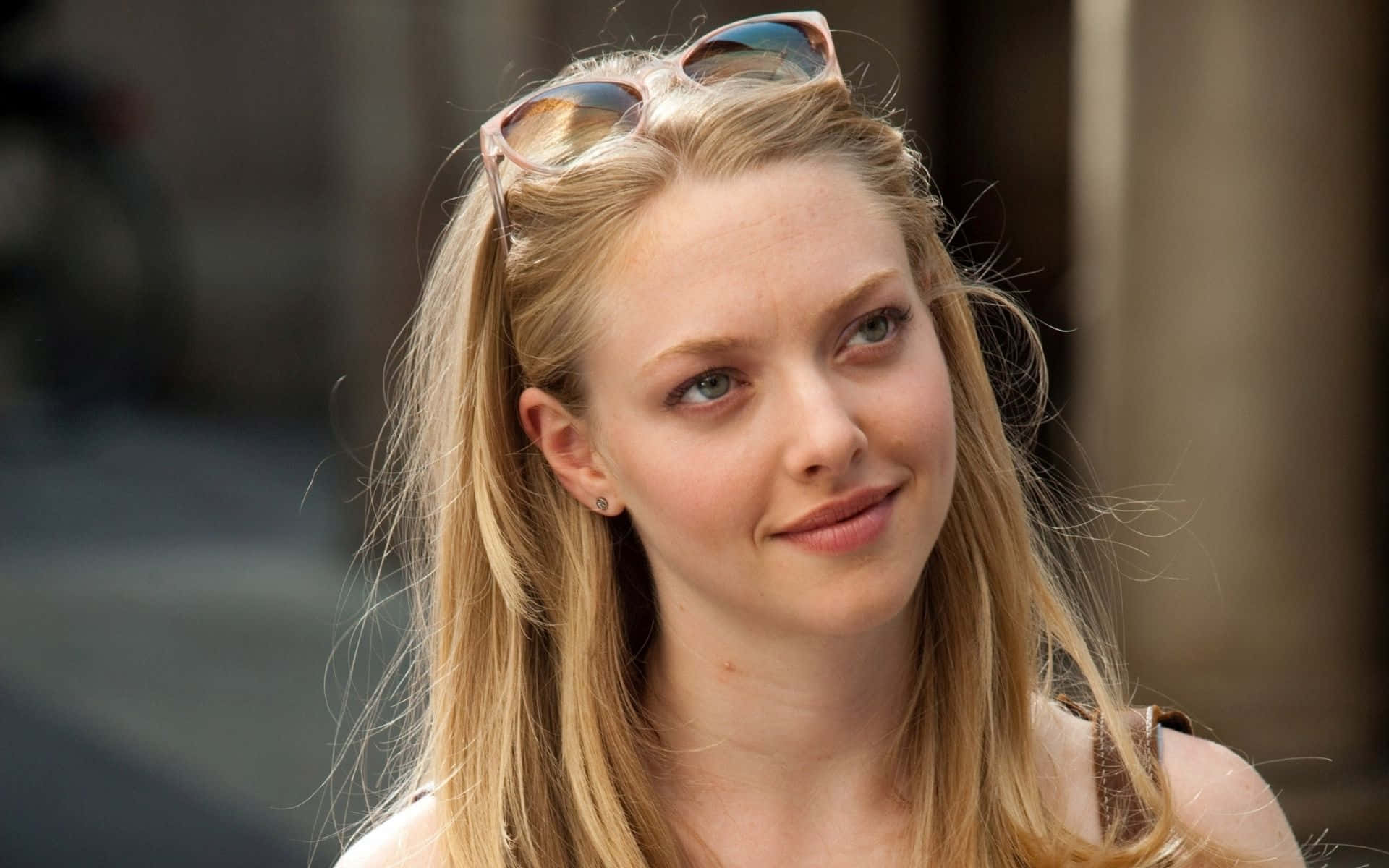 Amanda Seyfried 桌布 壁紙 — 一位金髮年輕女性，頭頂上架著太陽眼鏡，在城市環境中微笑著望向鏡頭外。
