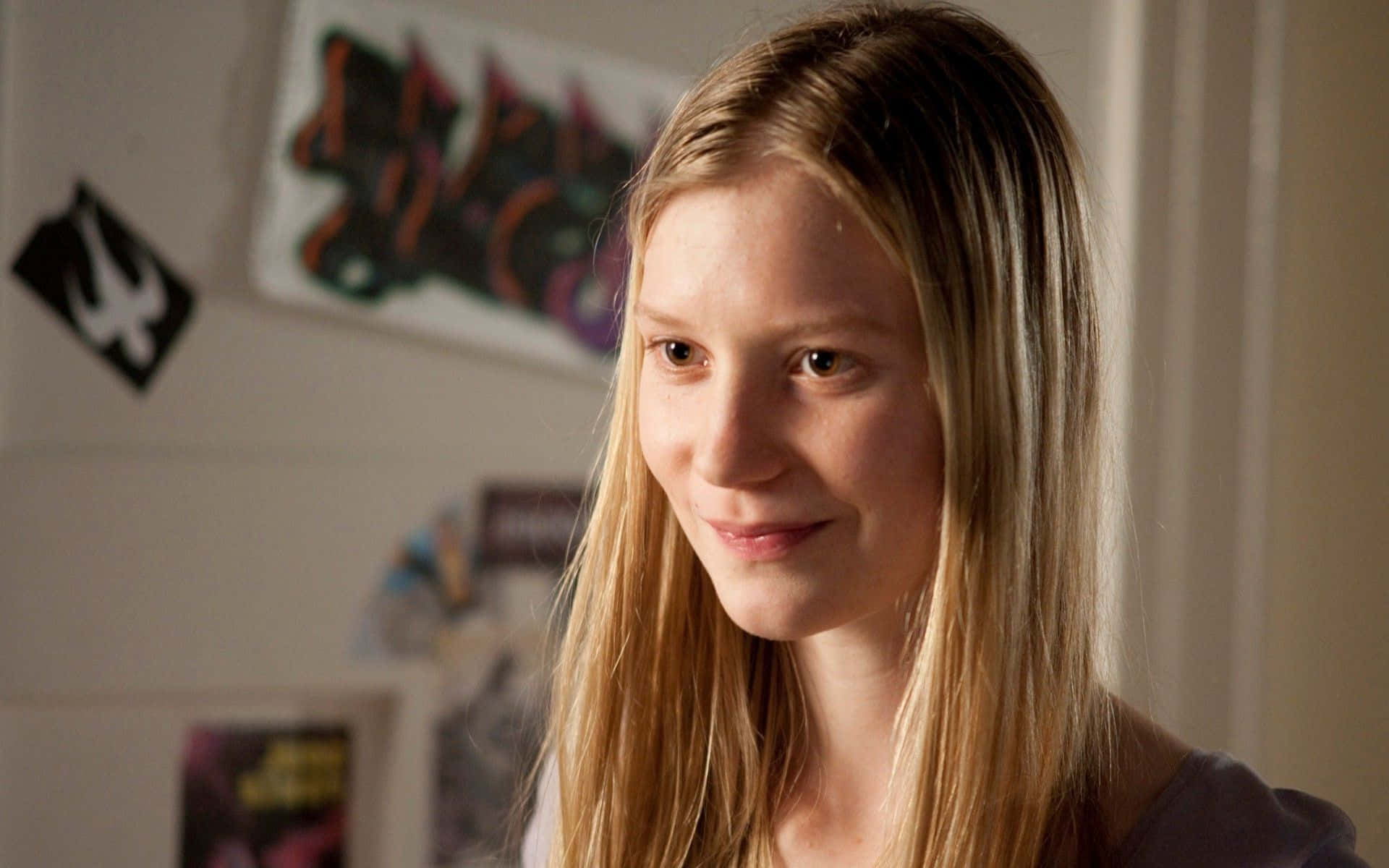 Mia Wasikowska 桌布 壁紙 — 一位金髮年輕女性在裝飾有塗鴉藝術和海報的房間裡溫柔地微笑。